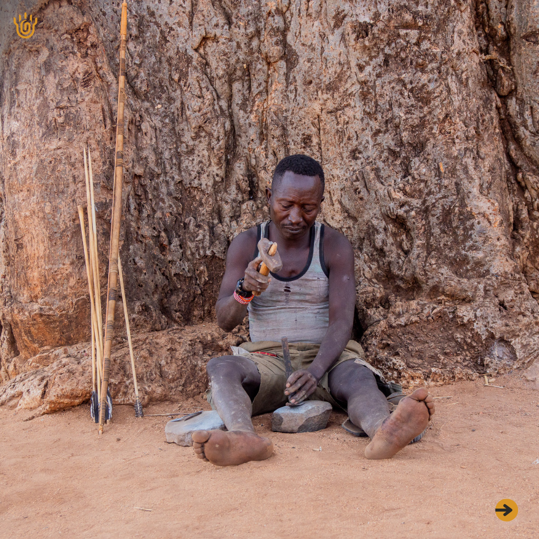 A picture showing a Hadzabe man sitting under a tree hammering at something whi