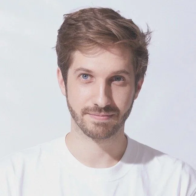 Portrait d'un jeune homme avec des cheveux bruns et une barbe légère, portant un t-shirt blanc, devant un fond clair.