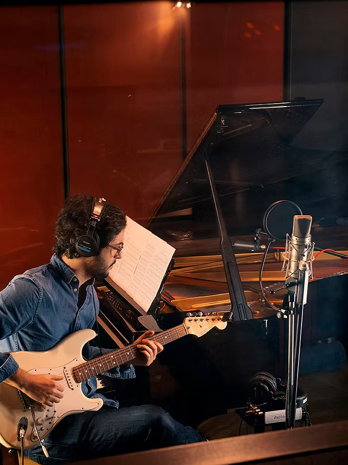 Un homme joue de la guitare électrique en studio d'enregistrement, avec un piano à queue en arrière-plan, portant des écouteurs et une chemise en denim, dans une ambiance chaleureuse et intimiste.