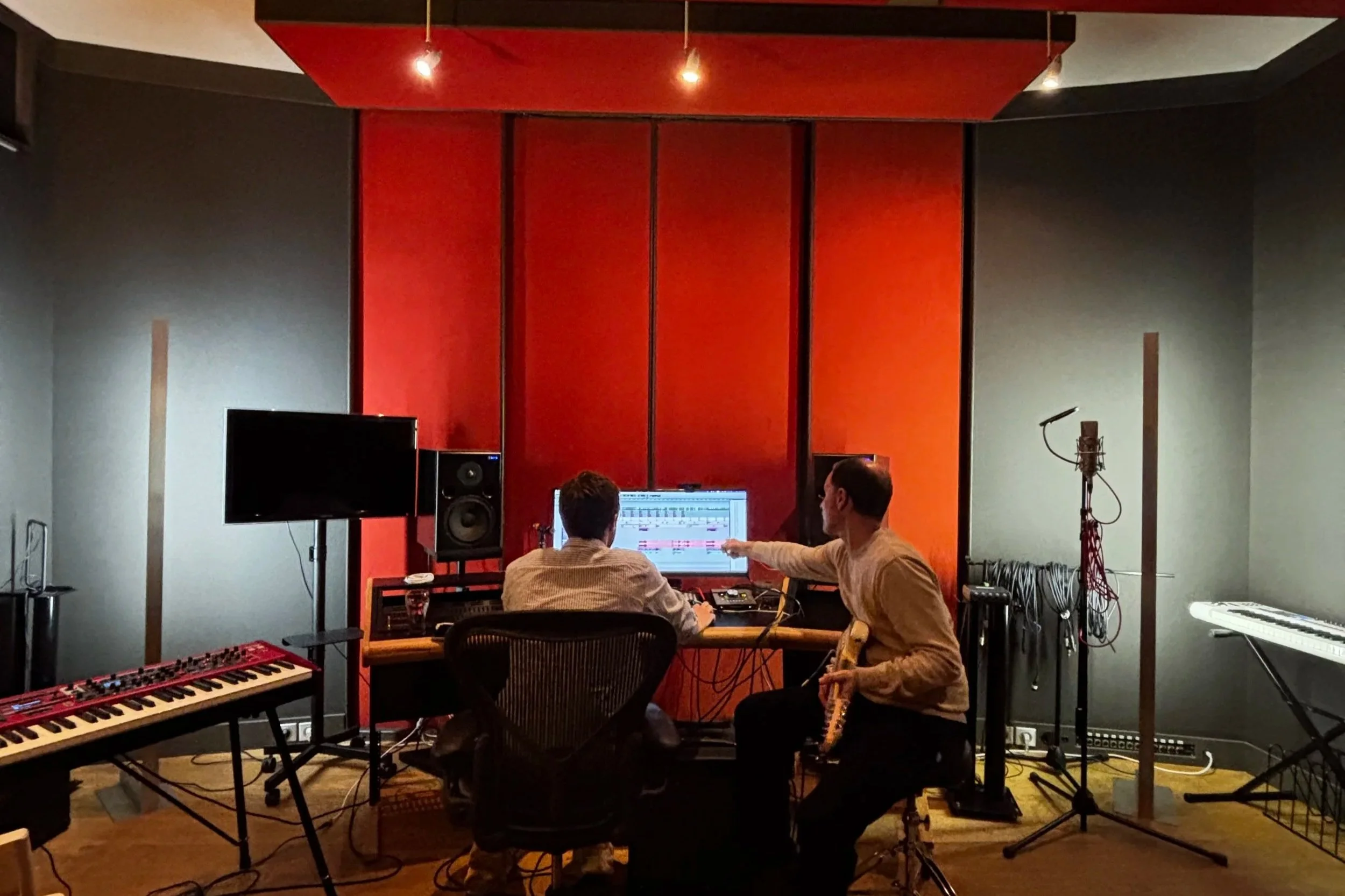 Deux hommes dans un studio d'enregistrement de musique avec divers équipements comme un clavier, une télévision, des haut-parleurs et un ordinateur, en train de discuter autour d'un écran.
