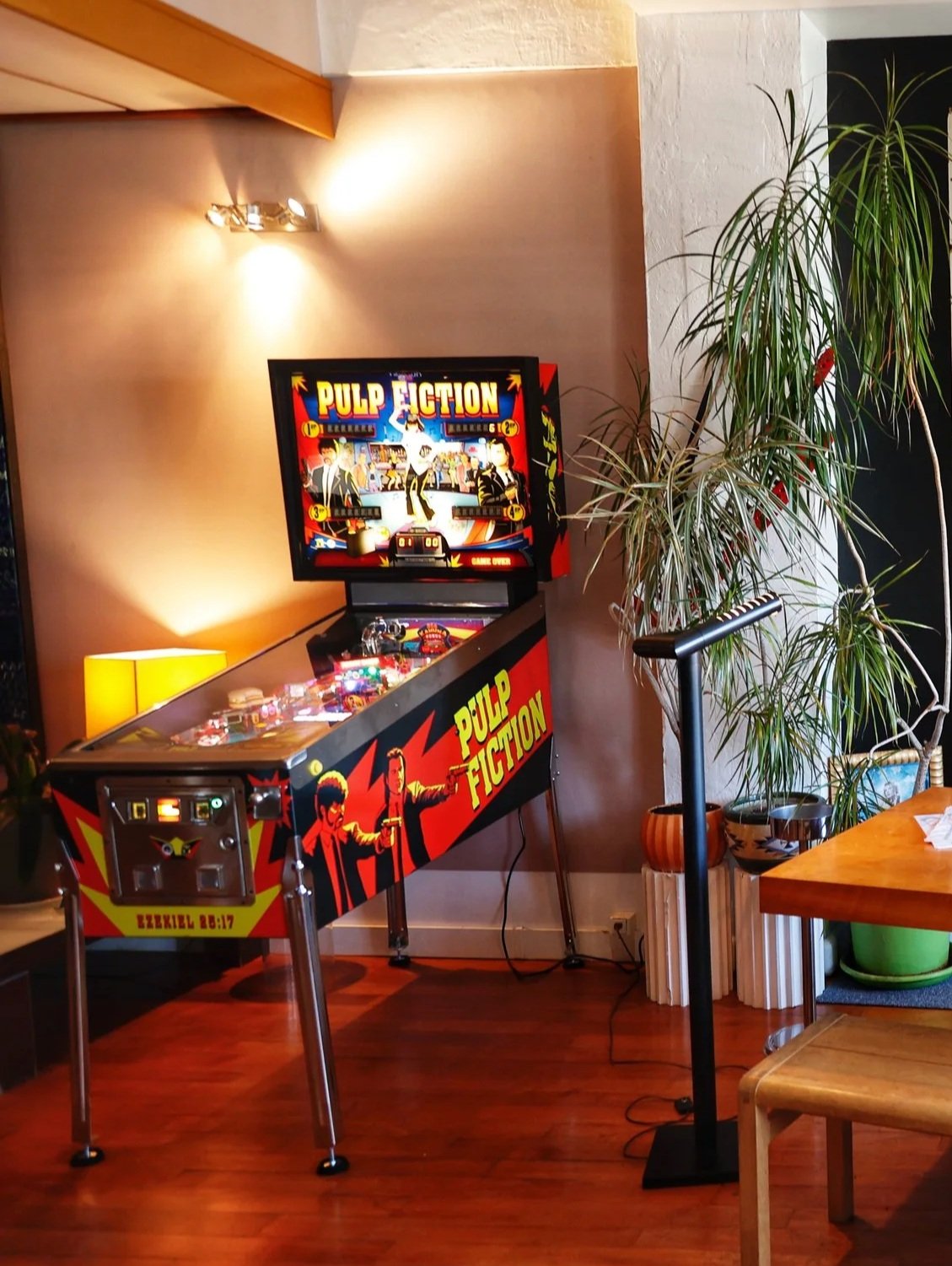 Une machine de jeu de flipper dans une pièce avec un décor rétro, une plante vert foncé à droite, une table en bois en premier plan, et un éclairage chaleureux.