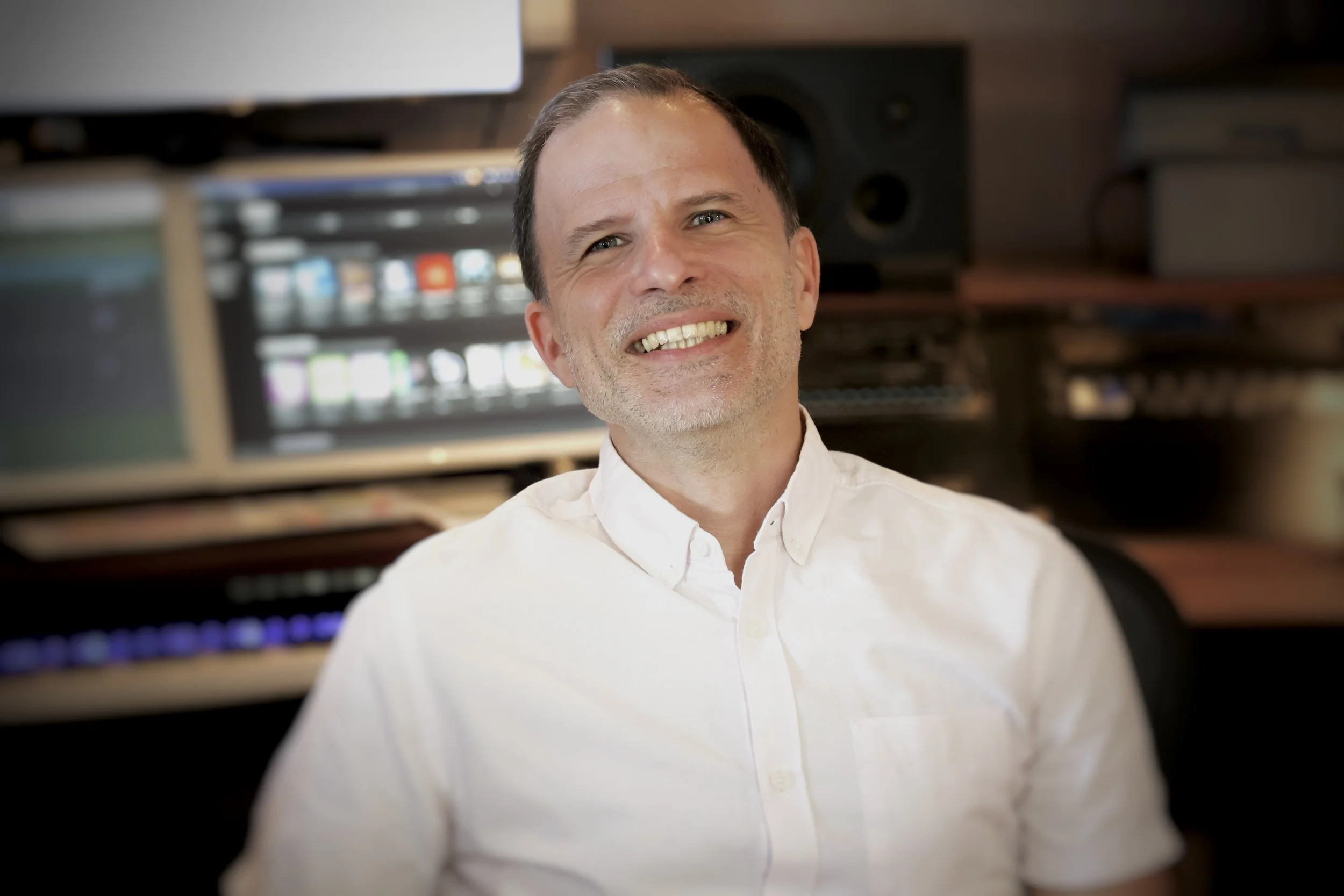 Un homme souriant en studio d'enregistrement ou salle de musique, portant une chemise blanche.