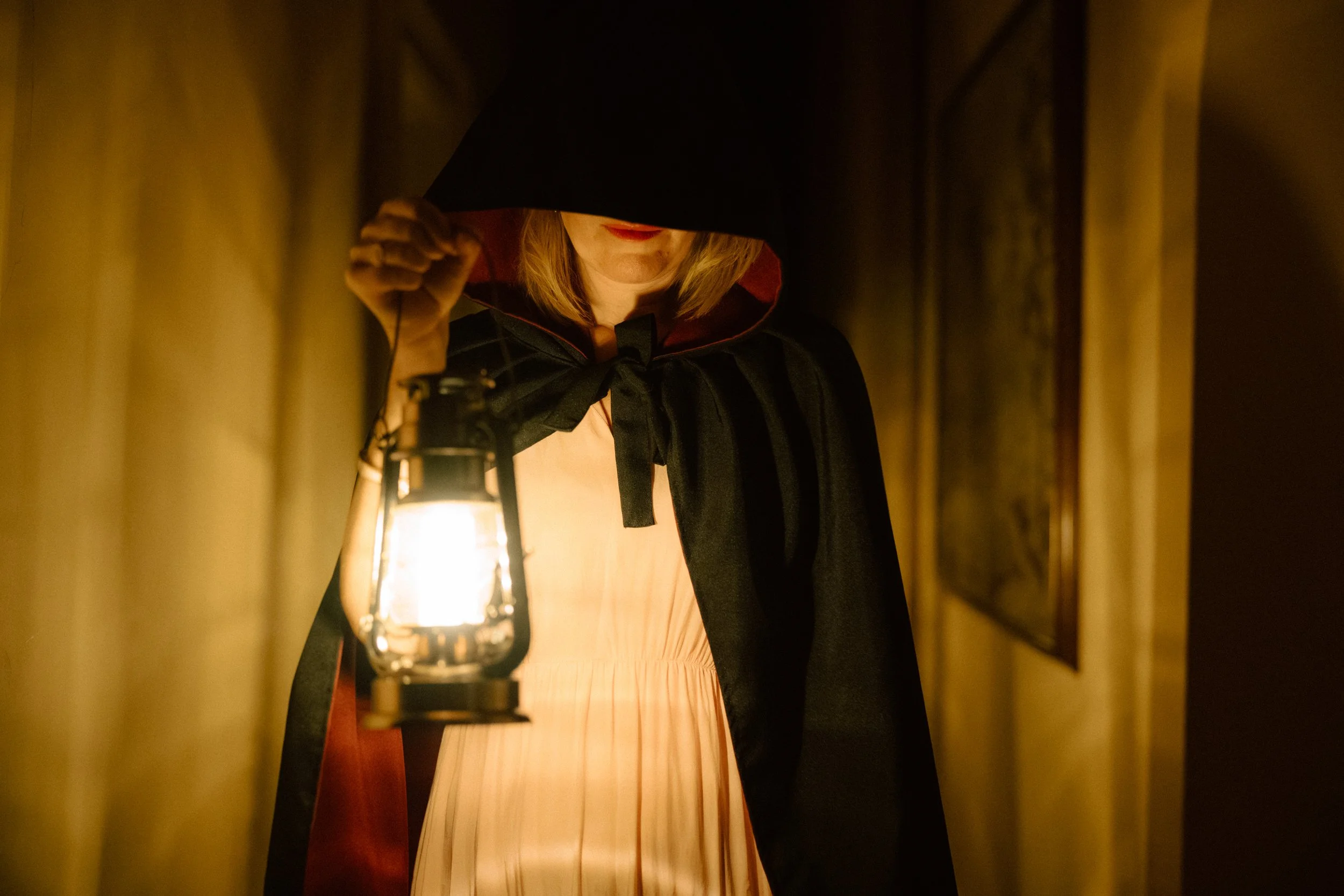A woman in a dark cloak holding a lantern in a dimly lit corridor, with her face partially obscured by the hood.