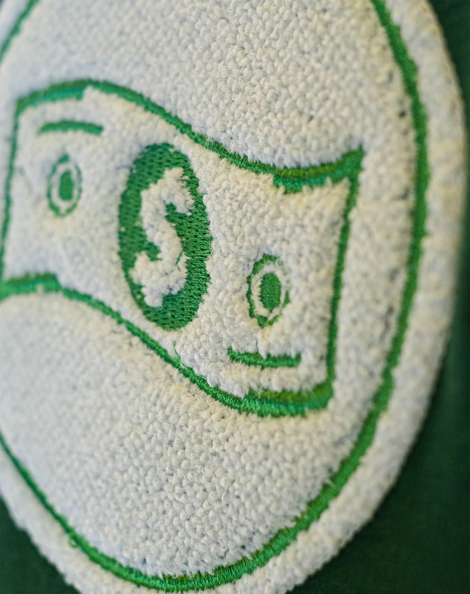 Nahaufnahme eines Stoff-Emblems mit grüner Stickerei auf weißem Hintergrund, das eine stilisierte Tasse oder ein Becher-Design zeigt.