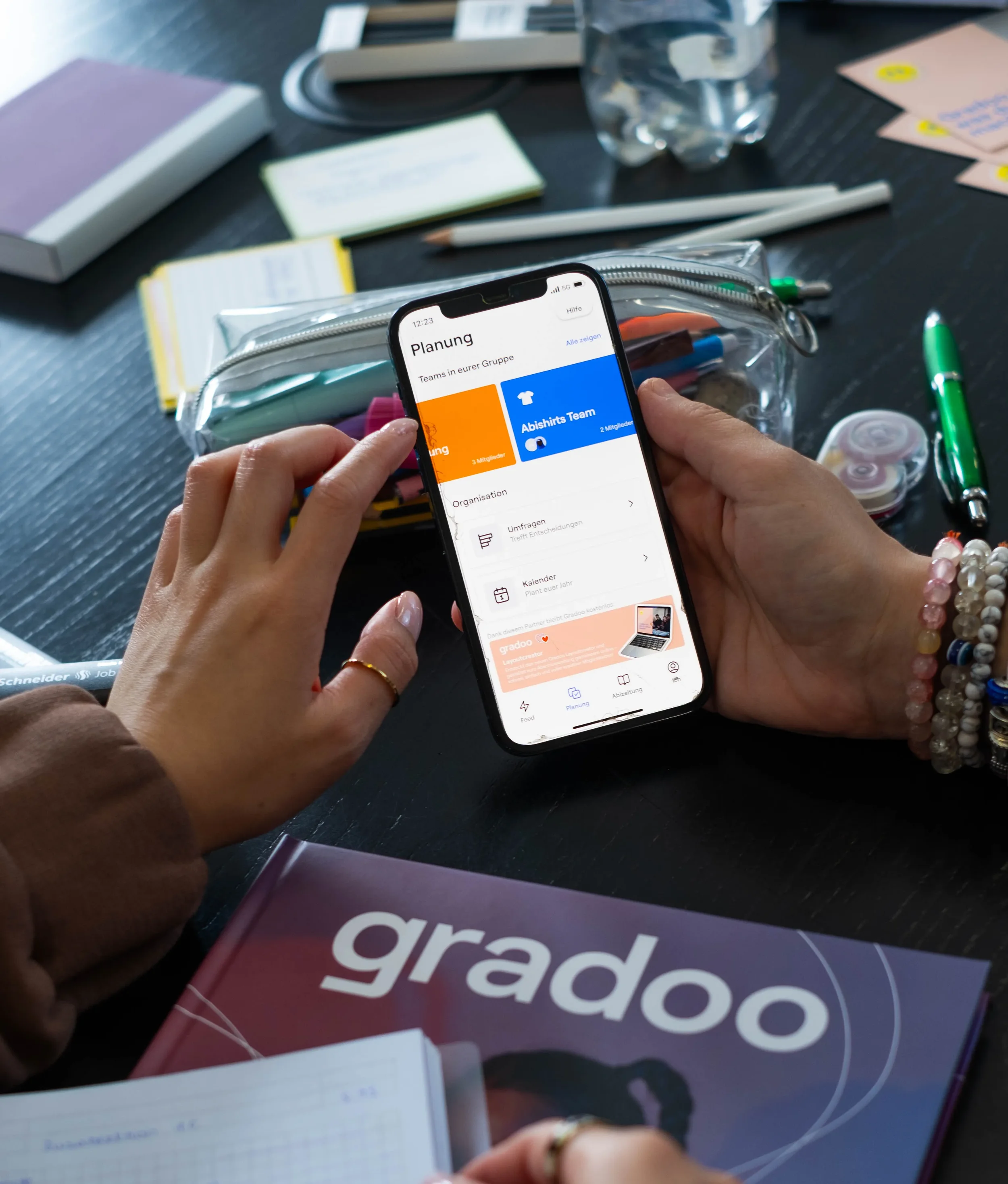 Nahaufnahme einer Person, die ein Smartphone hält, auf dem eine Planungs-App geöffnet ist, während sie an einem Schreibtisch mit Büchern, Stiften, Notizzetteln, einer Wasserflasche und einem Gradoo-Exemplar arbeitet.