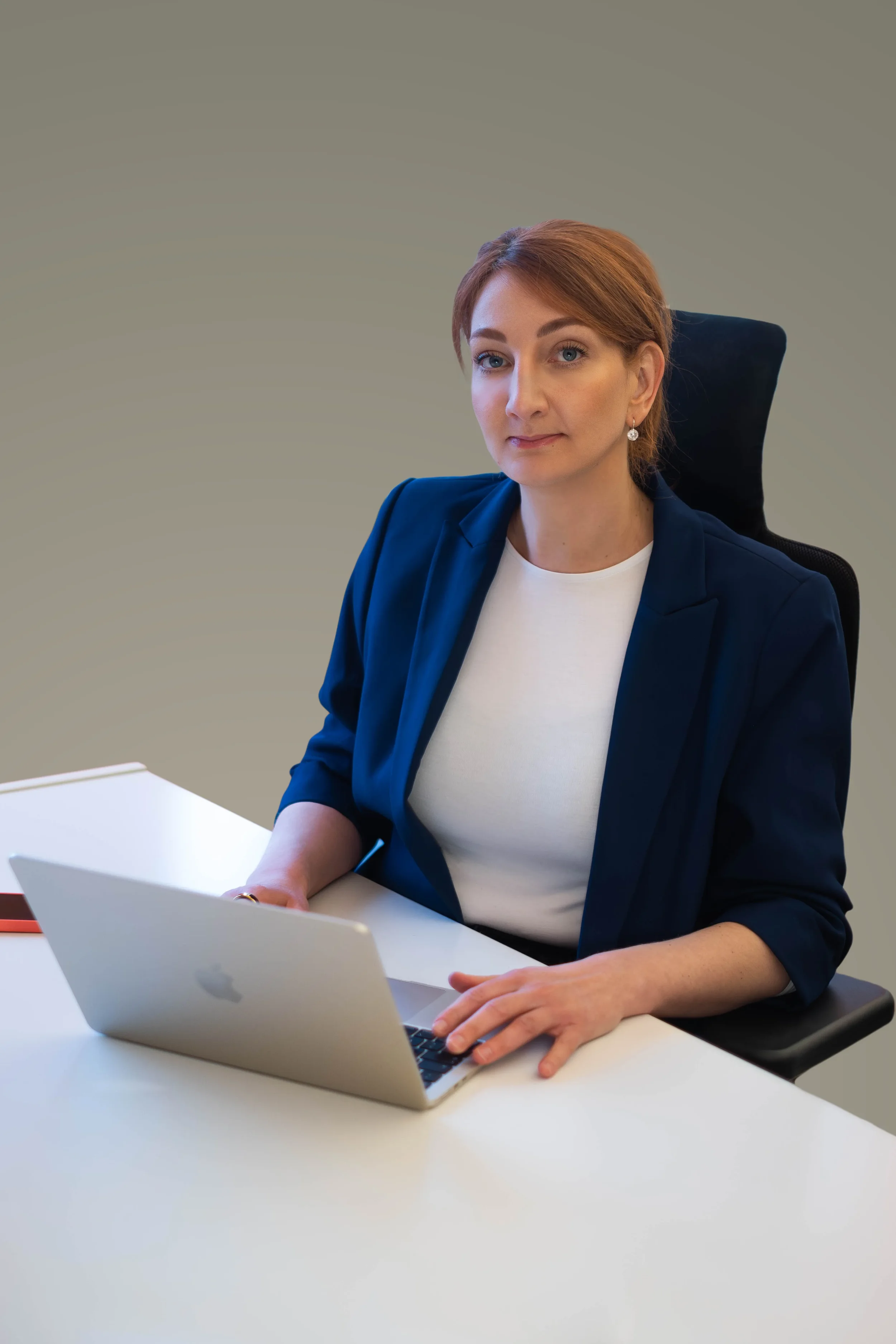 Eine Frau sitzt an einem weißen Schreibtisch mit einem offenen Laptop vor sich. Sie trägt einen dunkelblauen Blazer und eine weiße Bluse, hat rotbraunes Haar und trägt Ohrringe. Der Hintergrund ist neutral grau.