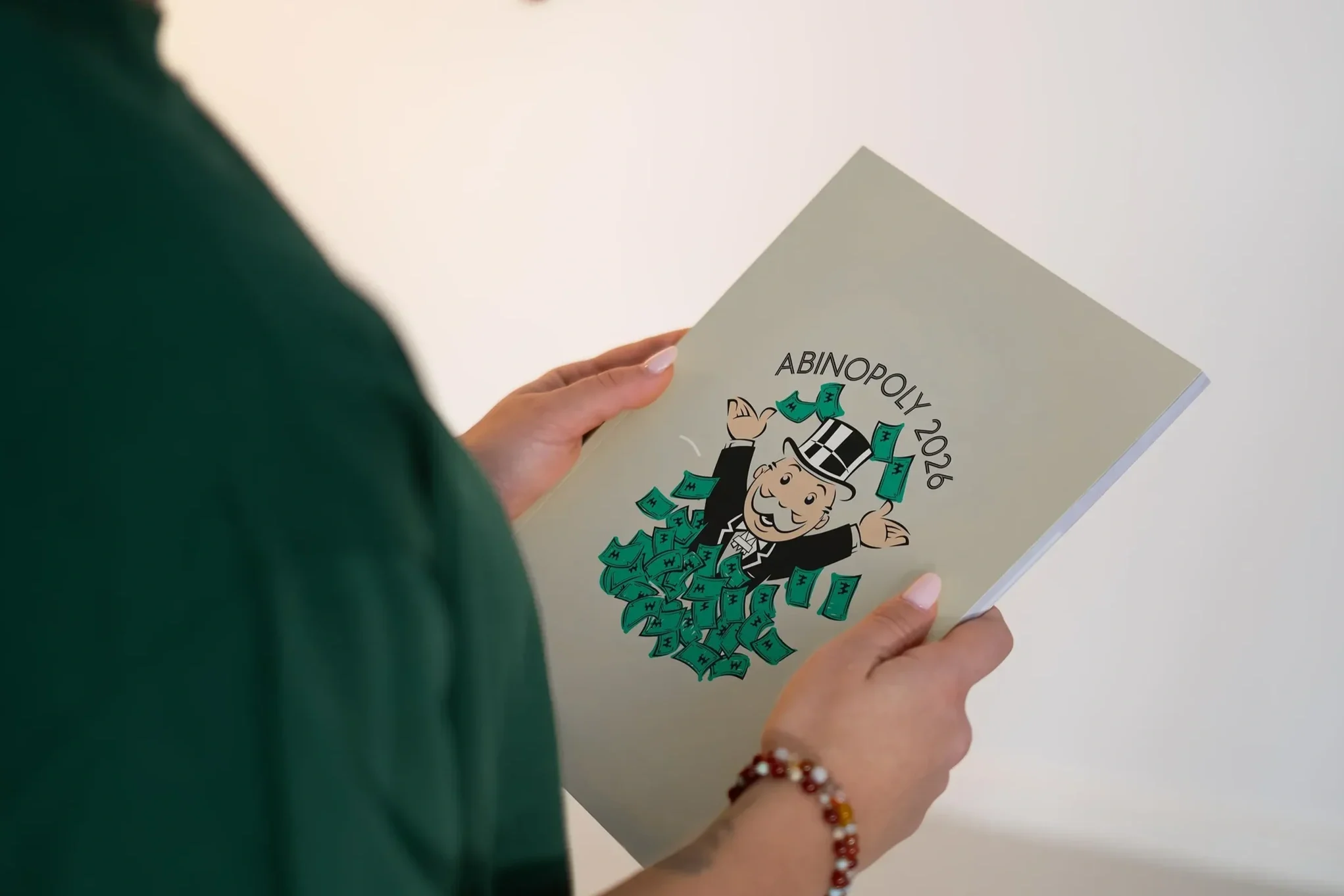 Eine Person hält ein Heft mit dem Titel "Abinopoly 2026". Auf dem Cover ist eine cartoonartige Figur eines Mannes in Anzug und Zylinderhut, der von Geldscheinen umgeben ist, dargestellt.