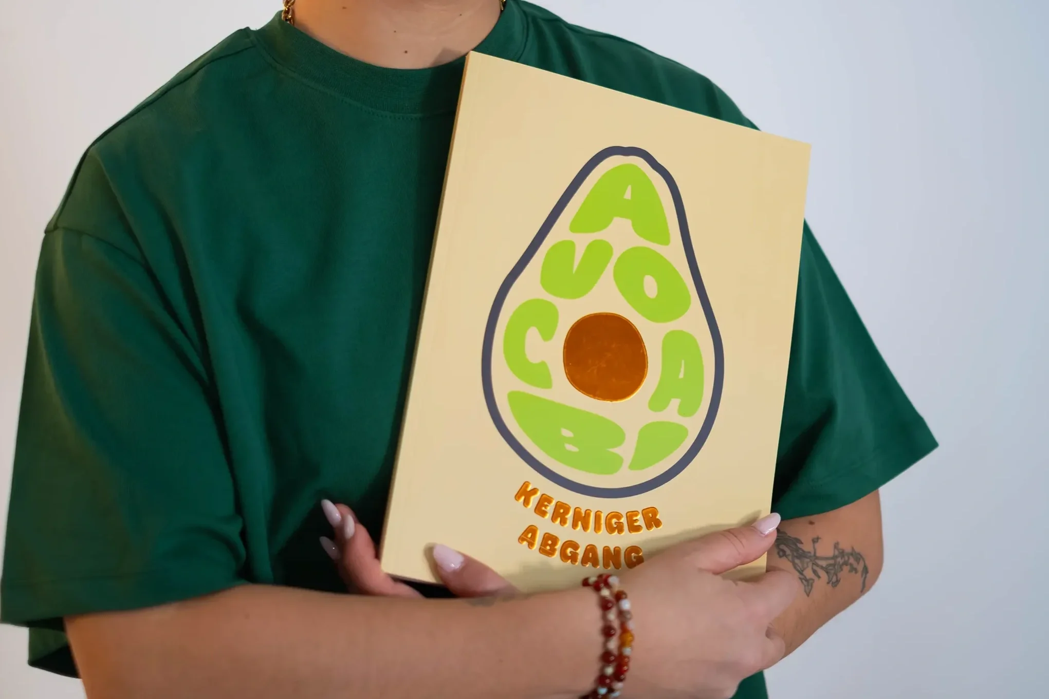 Person in grünes T-Shirt hält ein beige Buch mit einem Avocado-Design auf dem Cover, darunter steht 'KERNIGER ABGANG' in orange Buchstaben.