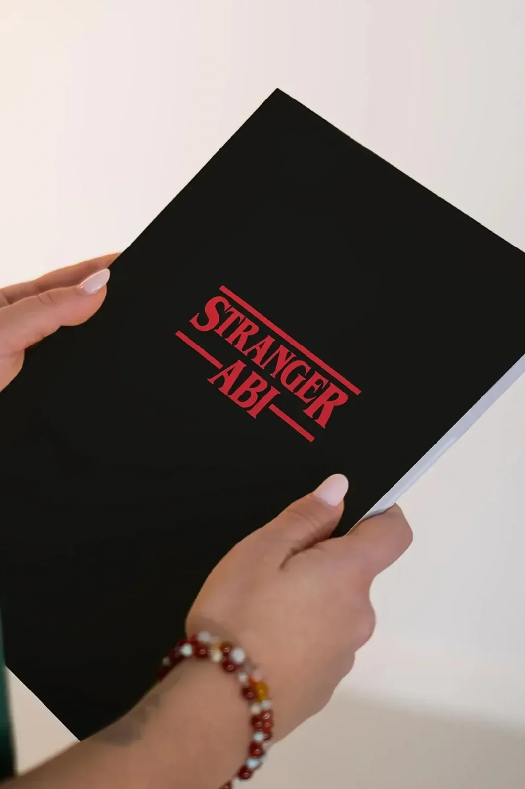 Hand hält ein schwarzes Buch mit rotem Schriftzug "STRANGER AB I" auf weißem Hintergrund.
