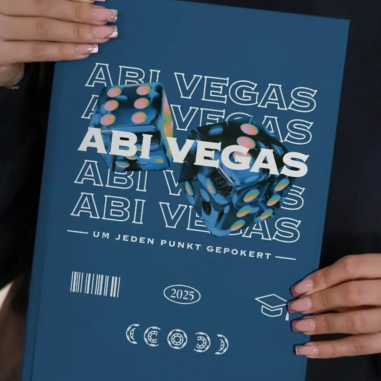 Person hält einen blauen Studienführer mit dem Titel 'ABI VEGAS' und zwei Würfel auf der Titelseite. Der Hintergrund ist dunkel, und die Person hat gepflegte Fingernägel.