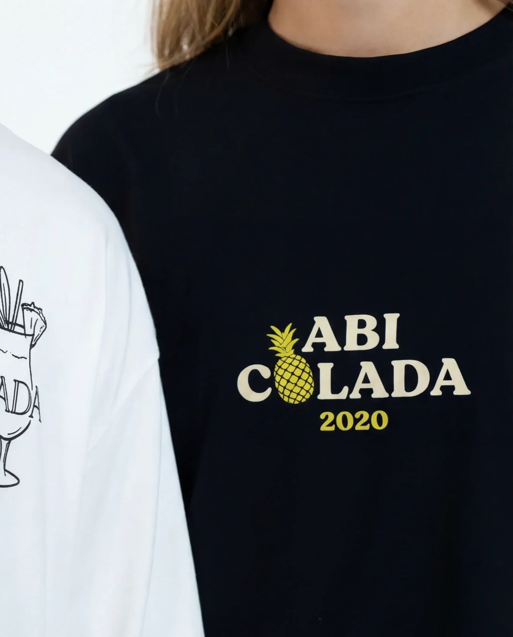 Zwei Personen tragen T-Shirts mit Grill- und Ananas-Designs, eines weiß mit einem Cocktailglas und dem Schriftzug 'ARGOLADA' und einem Hund, das andere schwarz mit einer Ananas und dem Text 'ABI COLADA 2020'.