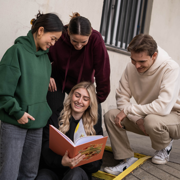 Gruppe von vier jungen Menschen, die draußen eine rote Abizeitung anschauen und lachen.