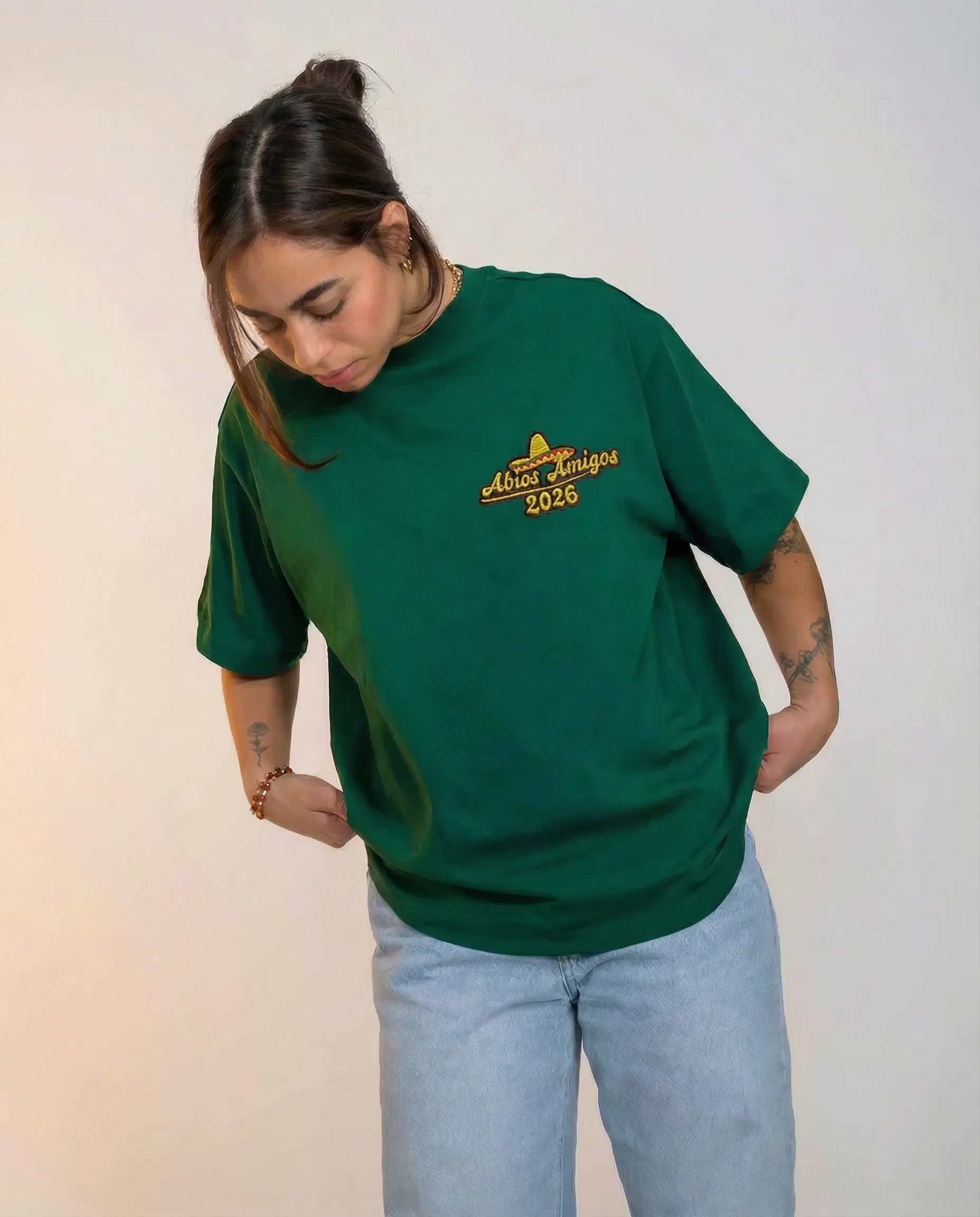 Junge Frau in grünem T-Shirt mit Aufdruck 'Abrazos Amigos 2026' und blauer Jeans, blickt nach unten, steht vor weißem Hintergrund.