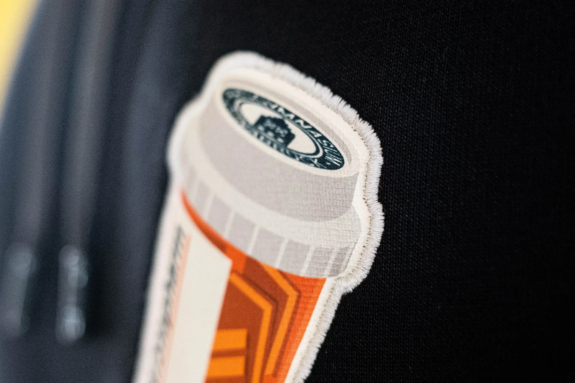 Detailaufnahme eines Stoffaufnähers auf schwarzem Textil, zeigt eine runde, mit Text und einem Logo versehene Designfläche.