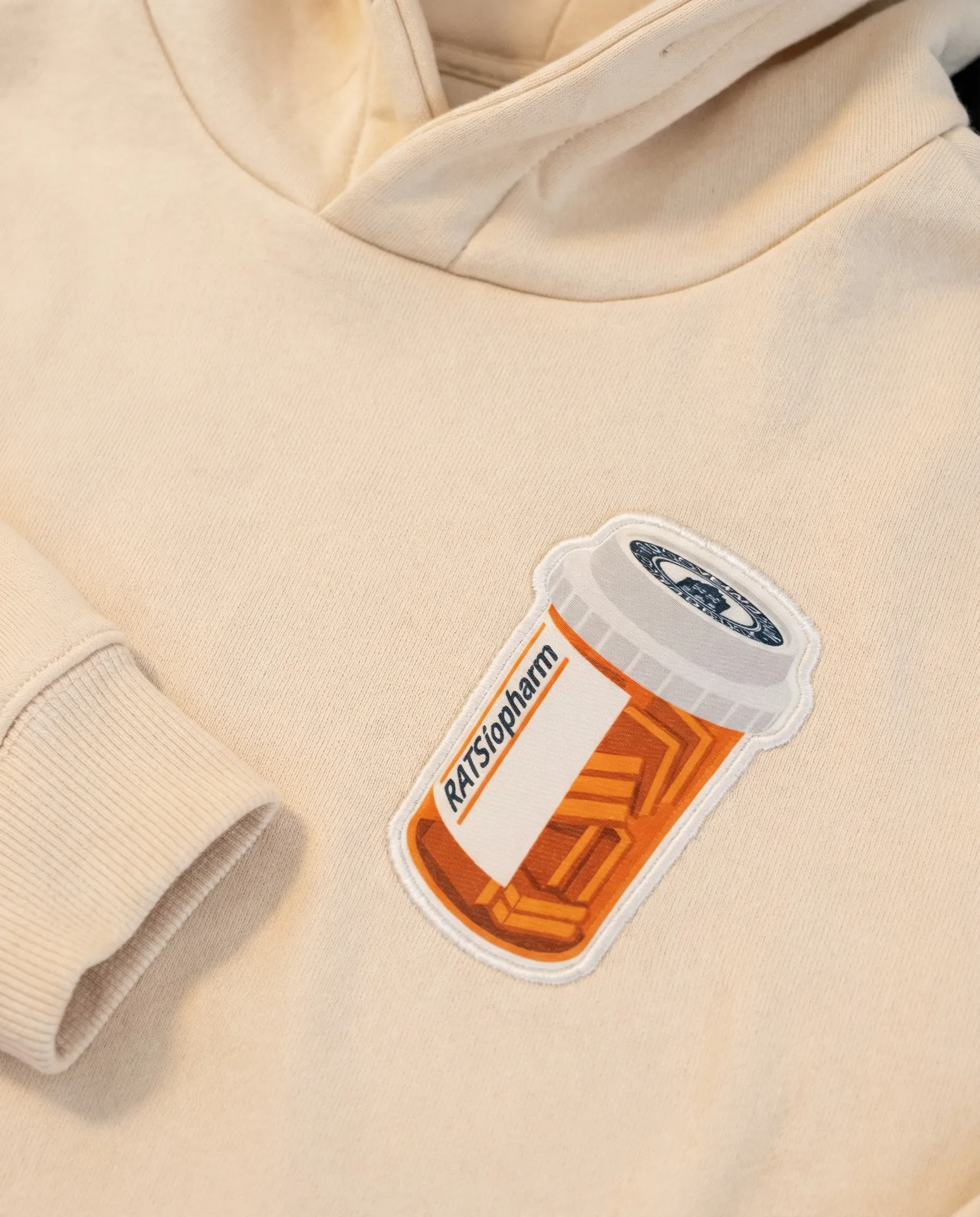 Naheaufnahme eines beige Kapuzenpullovers mit aufgenähtem Patch in Form einer Medikamentenpackung, auf der steht 'RATIOPHARM'.