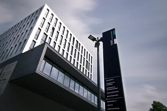 Modernes weißes und schwarzes Bürogebäude mit einem schwarzen Informationsschild und Straßenlaternen davor.