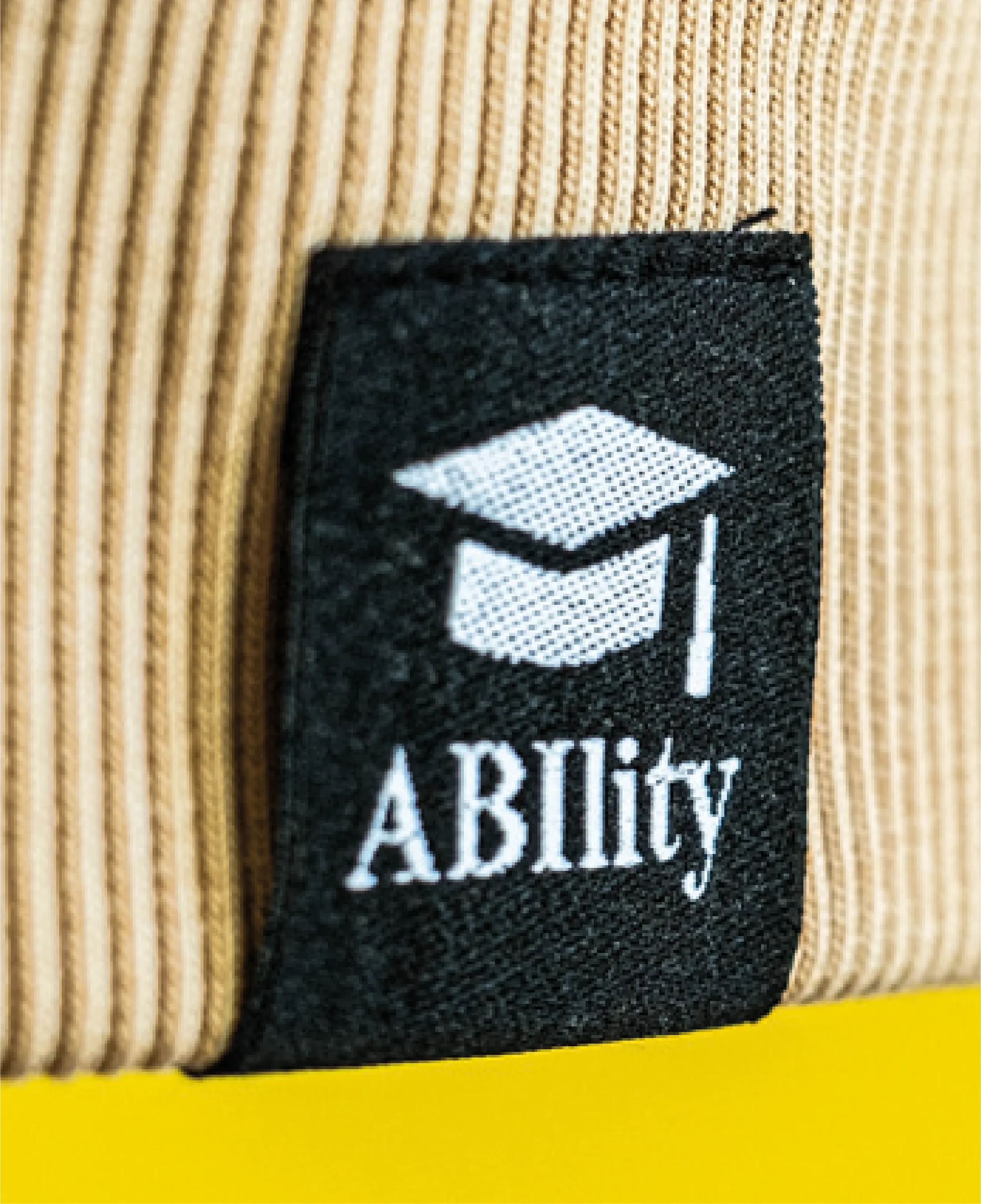Nähe eines beige gestreiften Stoffes mit einem schwarzen Etikett, das ein weißes Diplom- oder Hut-Symbol und den Text 'Ability' zeigt.