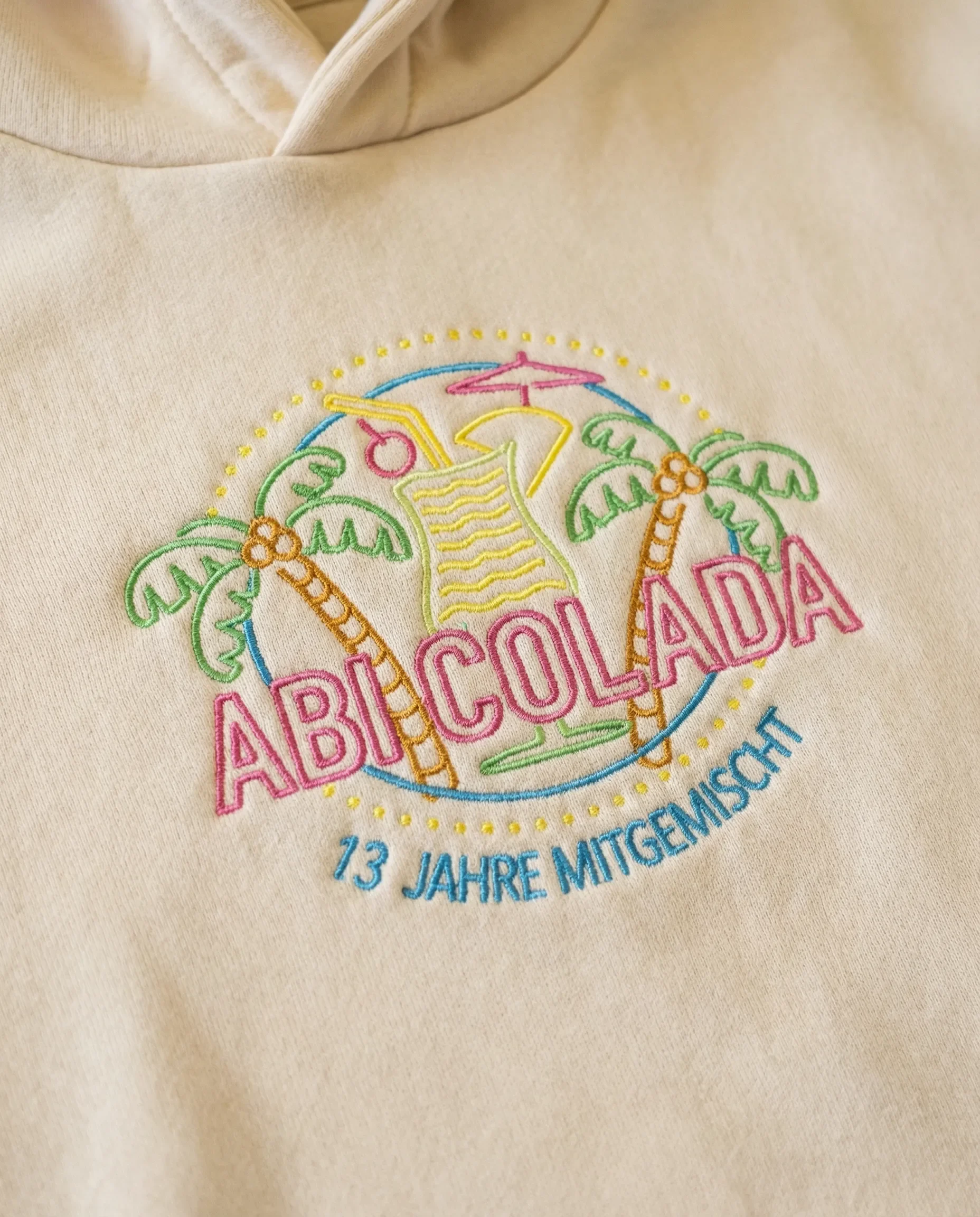 Aufgesticktes Design auf Kleidung mit Palmen, einem Cocktail, Sonnenschirm, und dem Text 'ABICOLADA' und '13 Jahre Mitgemacht'
