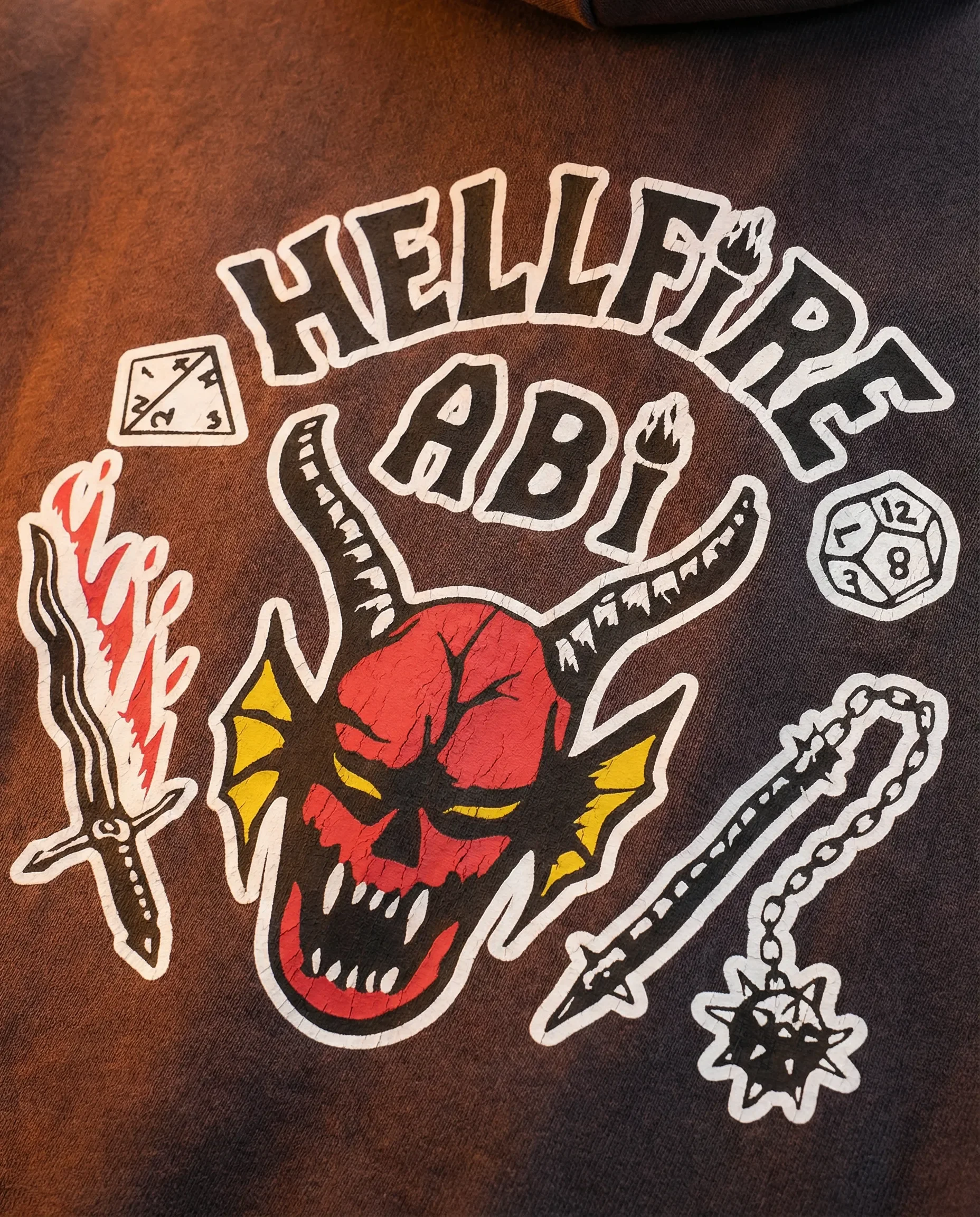 Ein schwarzes T-Shirt mit einem großen grafischen Druck, der einen roten Totenschädel mit schwarzen Kratzern und gelben Flügeln zeigt, umgeben von einem Messer, einer Kette mit einem Wurfstern, Würfeln und einem Pfeil.