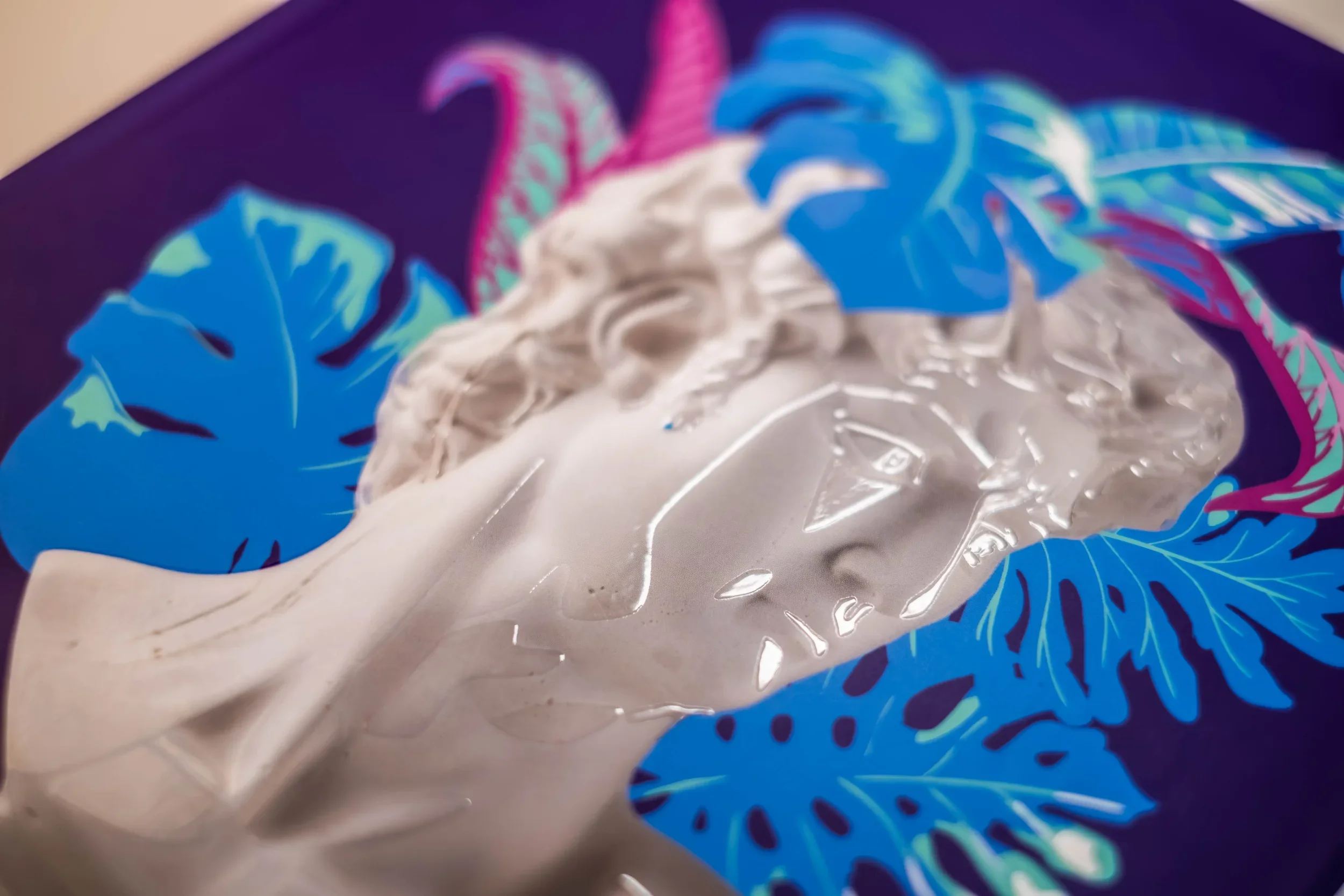 Nahaufnahme einer transparenten Skulptur eines Menschenkopfes vor einem Hintergrund mit blauen und pinkfarbenen tropischen Blättern.