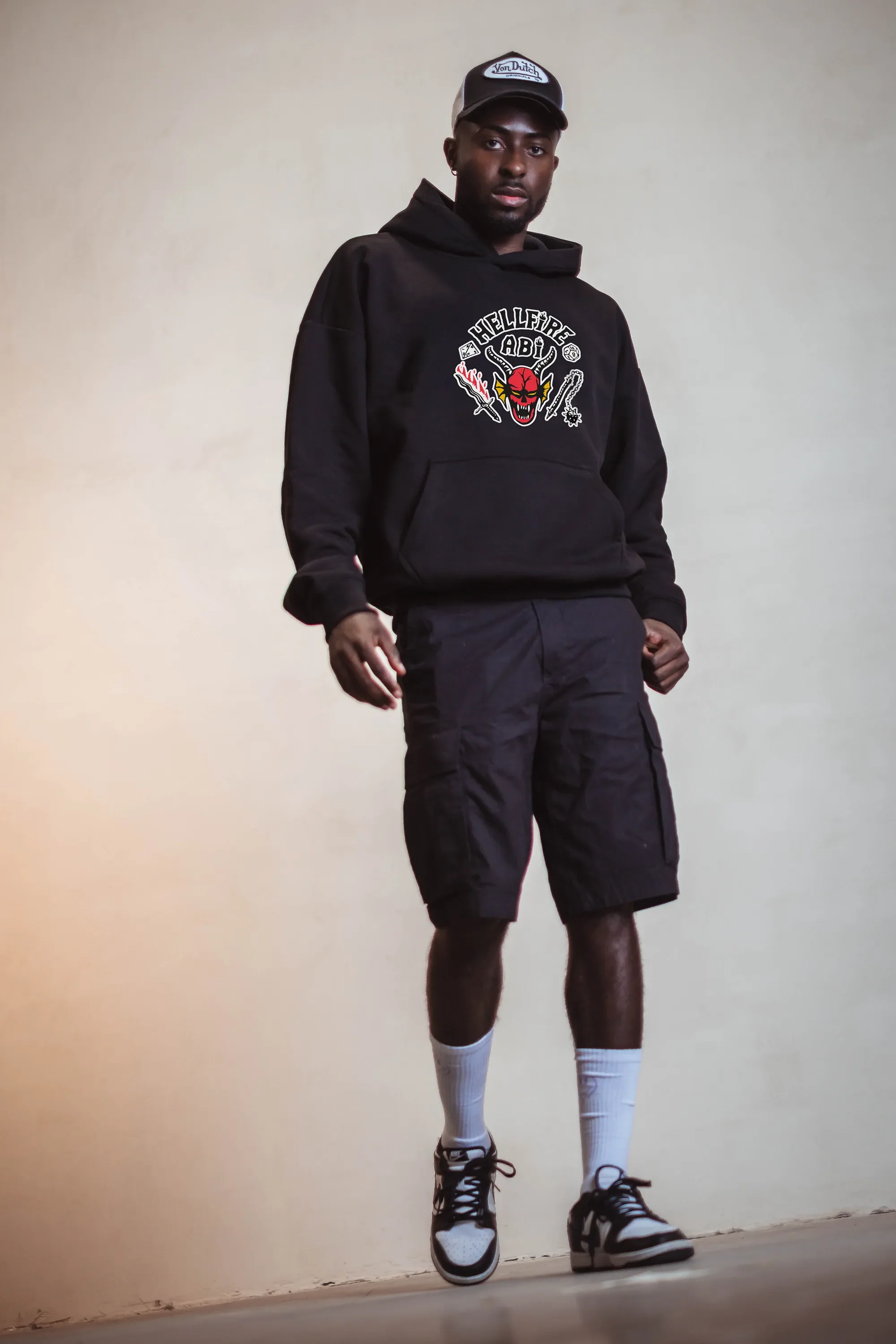 Ein junger Mann trägt eine schwarze Kapuzenjacke mit einem Totenkopf-Design und Schriftzug, schwarze Shorts, weiße Socken und Turnschuhe, steht vor einer beige Wand.