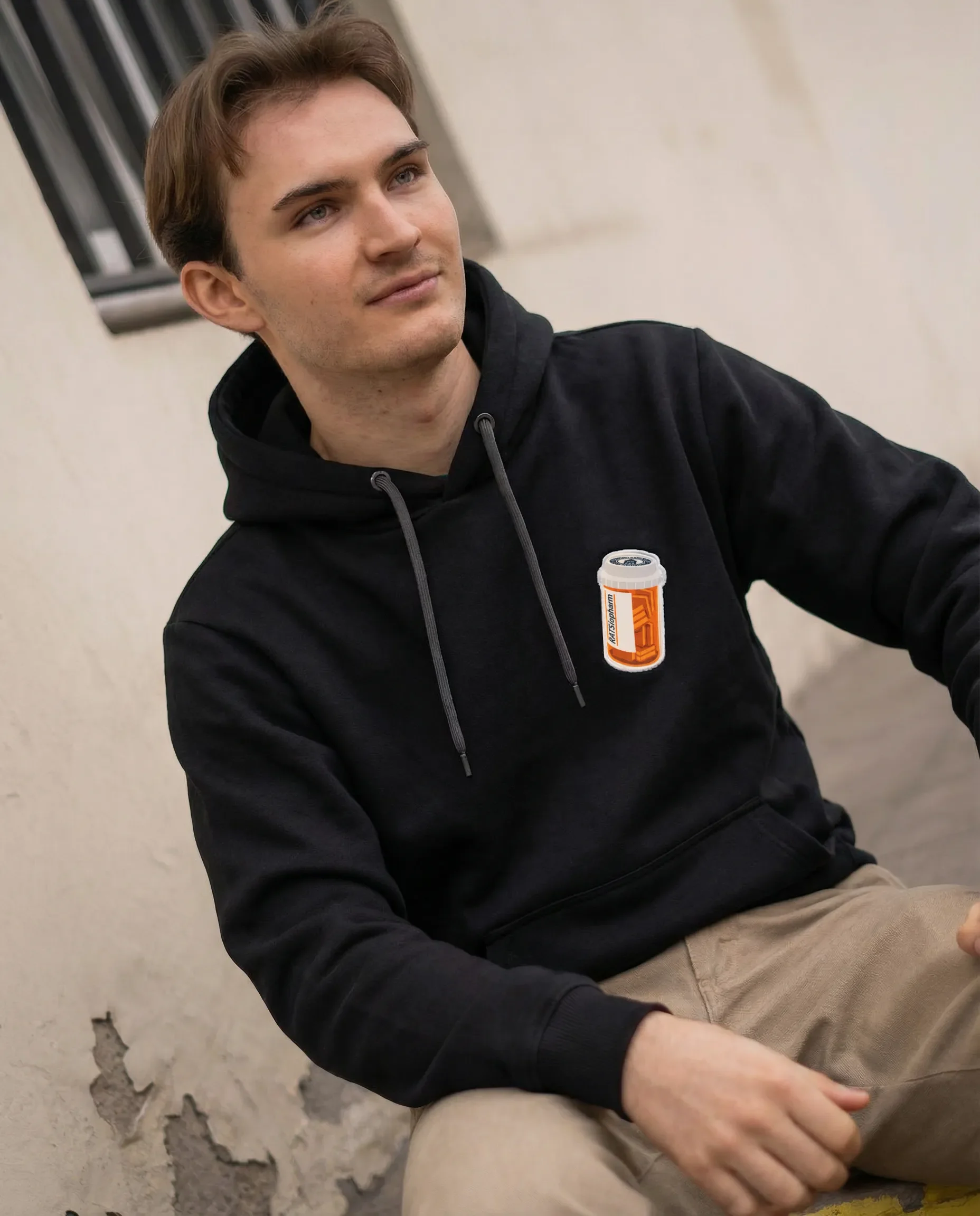 Junge trägt einen schwarzen Hoodie mit einem Aufnäher eines Coffee-Getränks und sitzt vor einer beschädigten Wand.