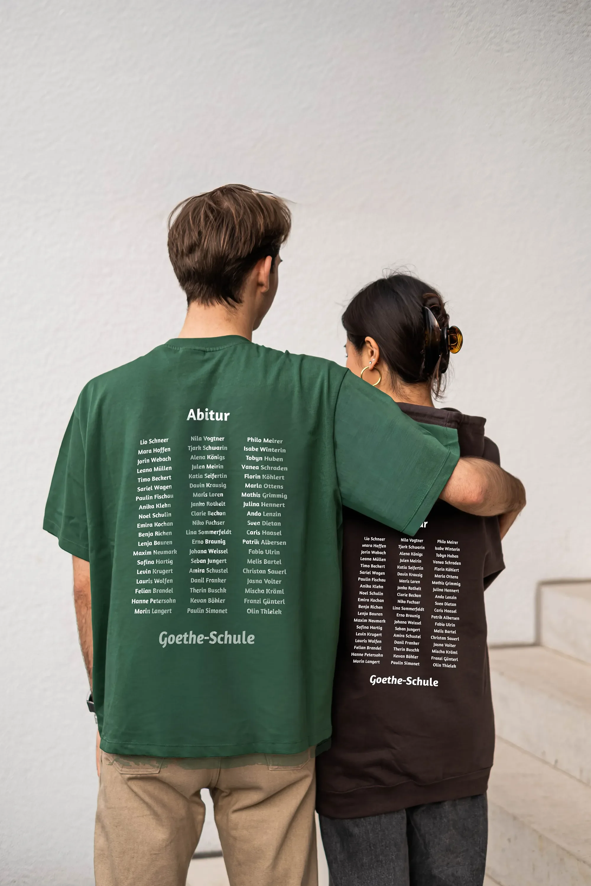 Zwei Personen stehen mit ihren Rücken zueinander, tragen grüne und braune T-Shirts mit einer Liste von Namen und der Aufschrift 'Abitur' sowie 'Goethe-Schule' auf der Rückseite.