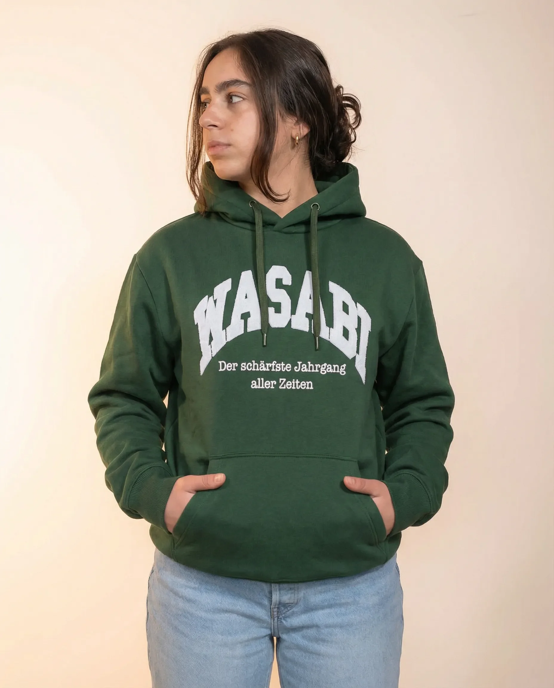 Junge Frau trägt grüne Kapuzenjacke mit weißen Buchstaben und deutschem Text, Jeans, steht vor heller Wand.
