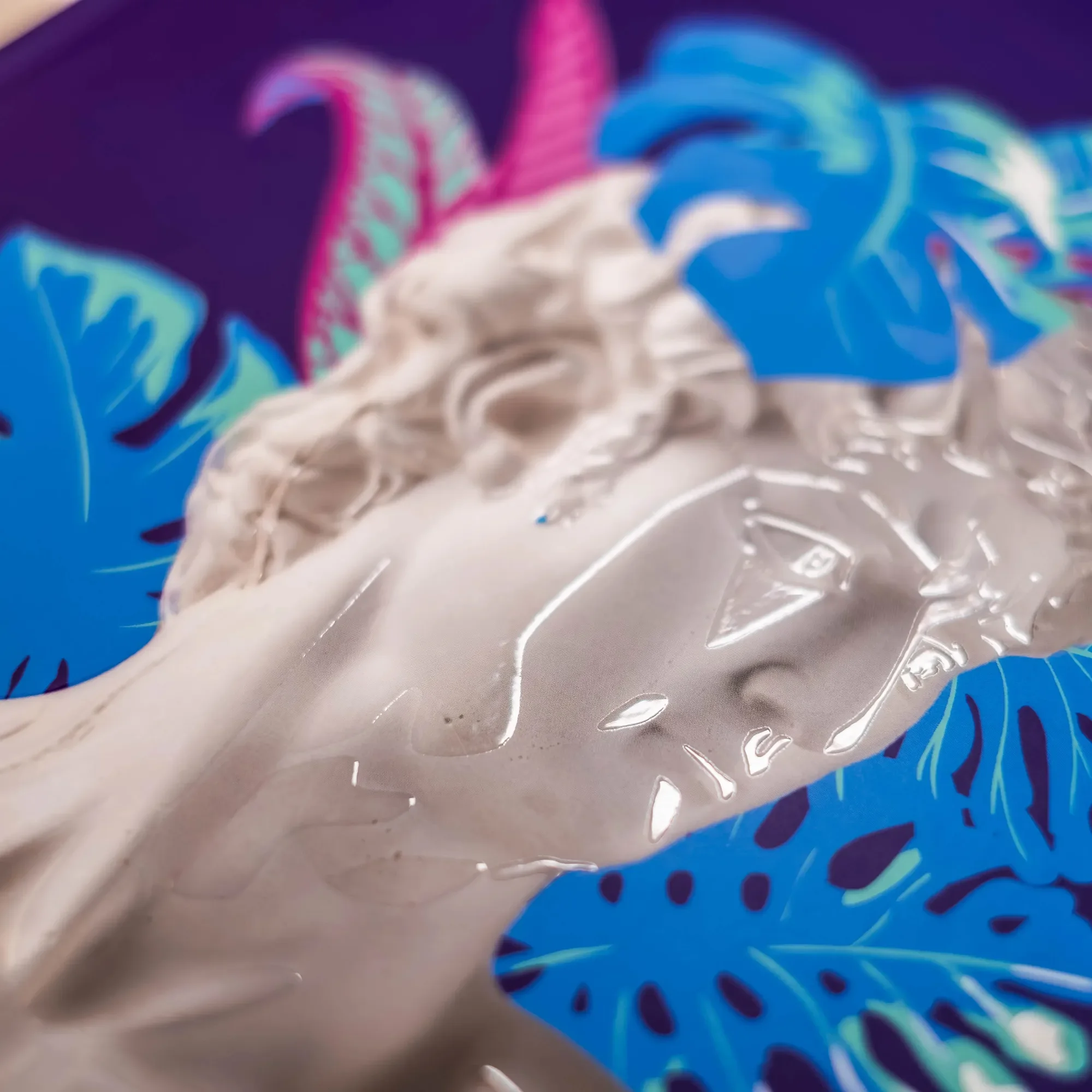 Nahaufnahme einer transparenten Skulptur eines Menschenkopfes vor einem Hintergrund mit blauen und pinkfarbenen tropischen Blättern.