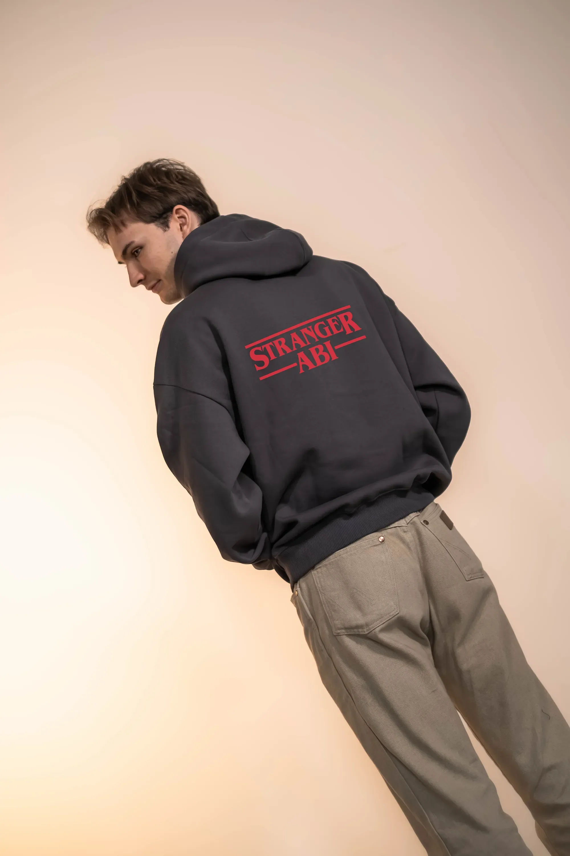 Junger Mann trägt grauen Hosen und schwarze Hoodie mit rotem Text "Stranger Ab" auf dem Rücken, vor hellem Hintergrund.