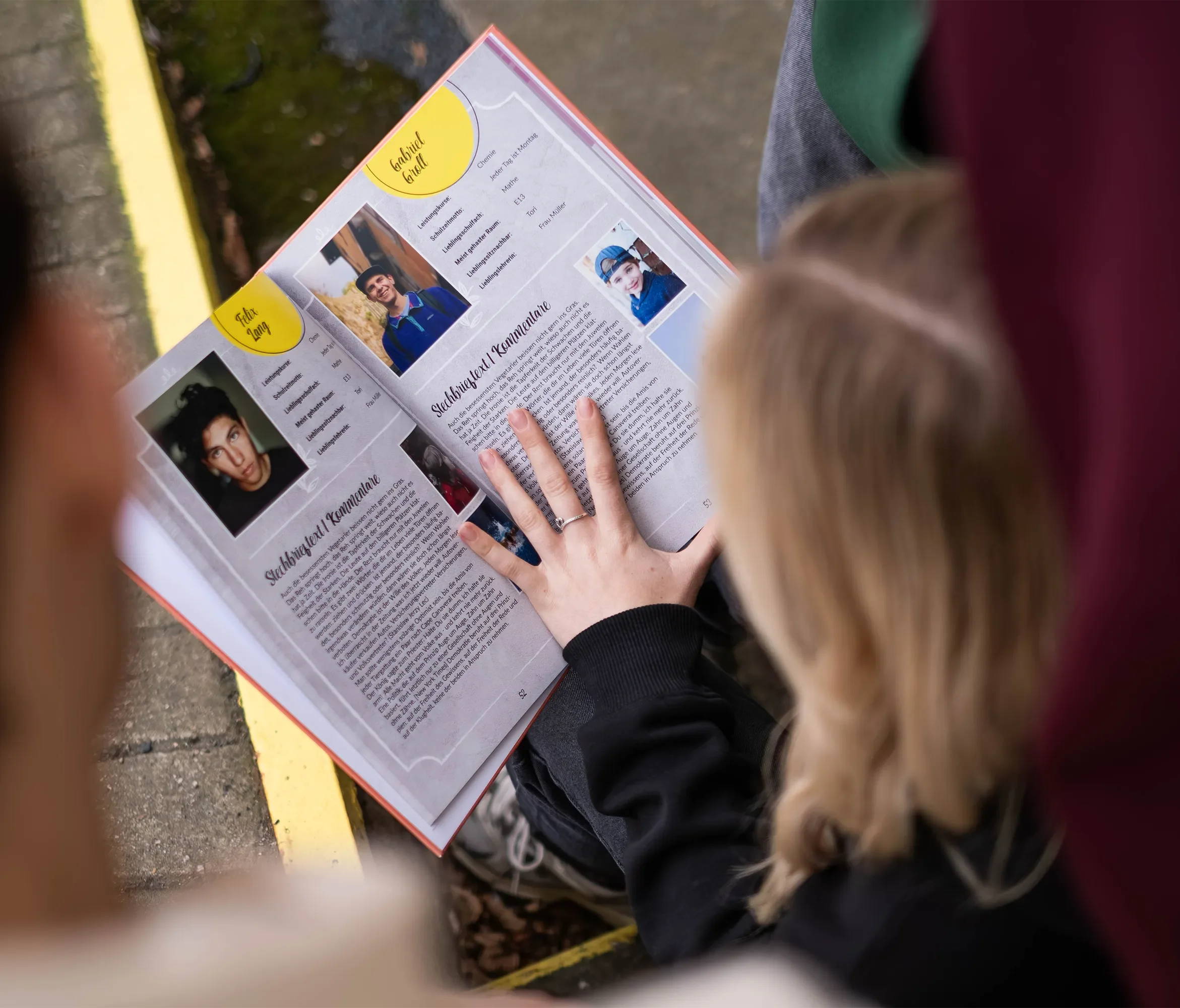 Zwei Personen lesen eine Broschüre mit Fotos und Texten, wahrscheinlich eine Schul- oder Vereinszeitung. Der Fokus liegt auf der Broschüre.