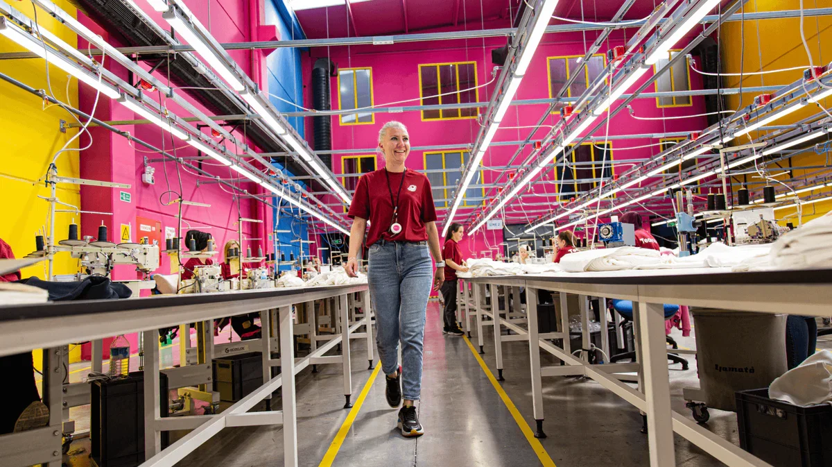 Mitarbeiterin in einem bunten industriellen Textilfabrik mit langen Arbeitstischen, Maschinen und farbigen Wänden, die pink, blau und gelb sind.