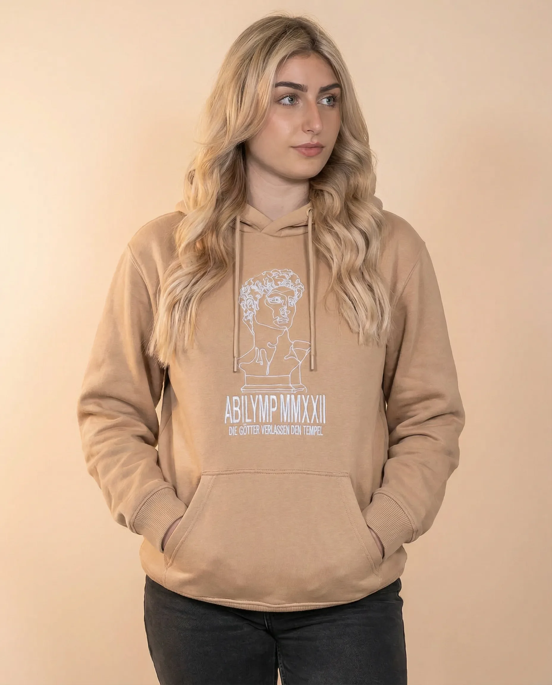 Junge Frau in beigem Hoodie mit antiker römischer Statue und lateinischer Schrift, die auf ein religiöses Ereignis hinweist, vor neutralem Hintergrund.