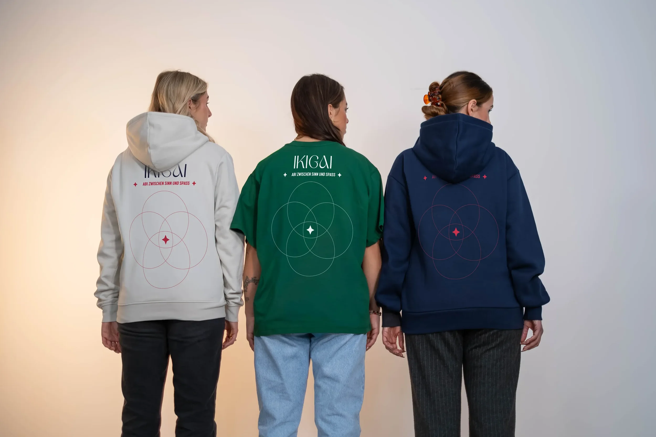 Drei Frauen stehen mit Rücken zur Kamera, tragen hoodies in Weiß, Grün und Blau, mit einer grafischen Gestaltung und Text auf dem Rücken, vor einem neutralen Hintergrund.