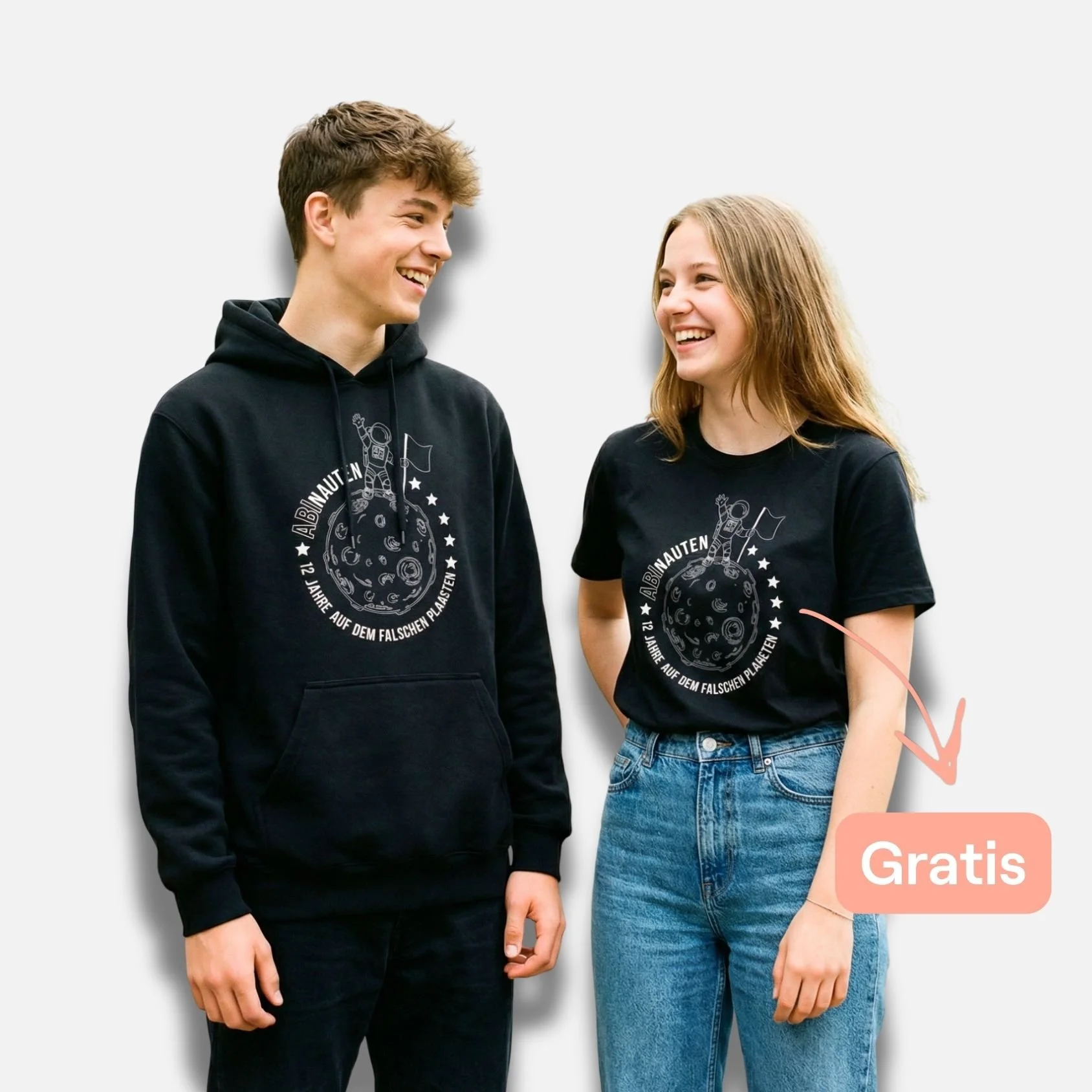 Zwei Jugendliche tragen schwarze T-Shirts mit einem Weltraummotiv und Text. Das Mädchen zeigt auf ein T-Shirt, auf dem steht 'Gratis'. Beide lachen. Hintergrund ist weiß.