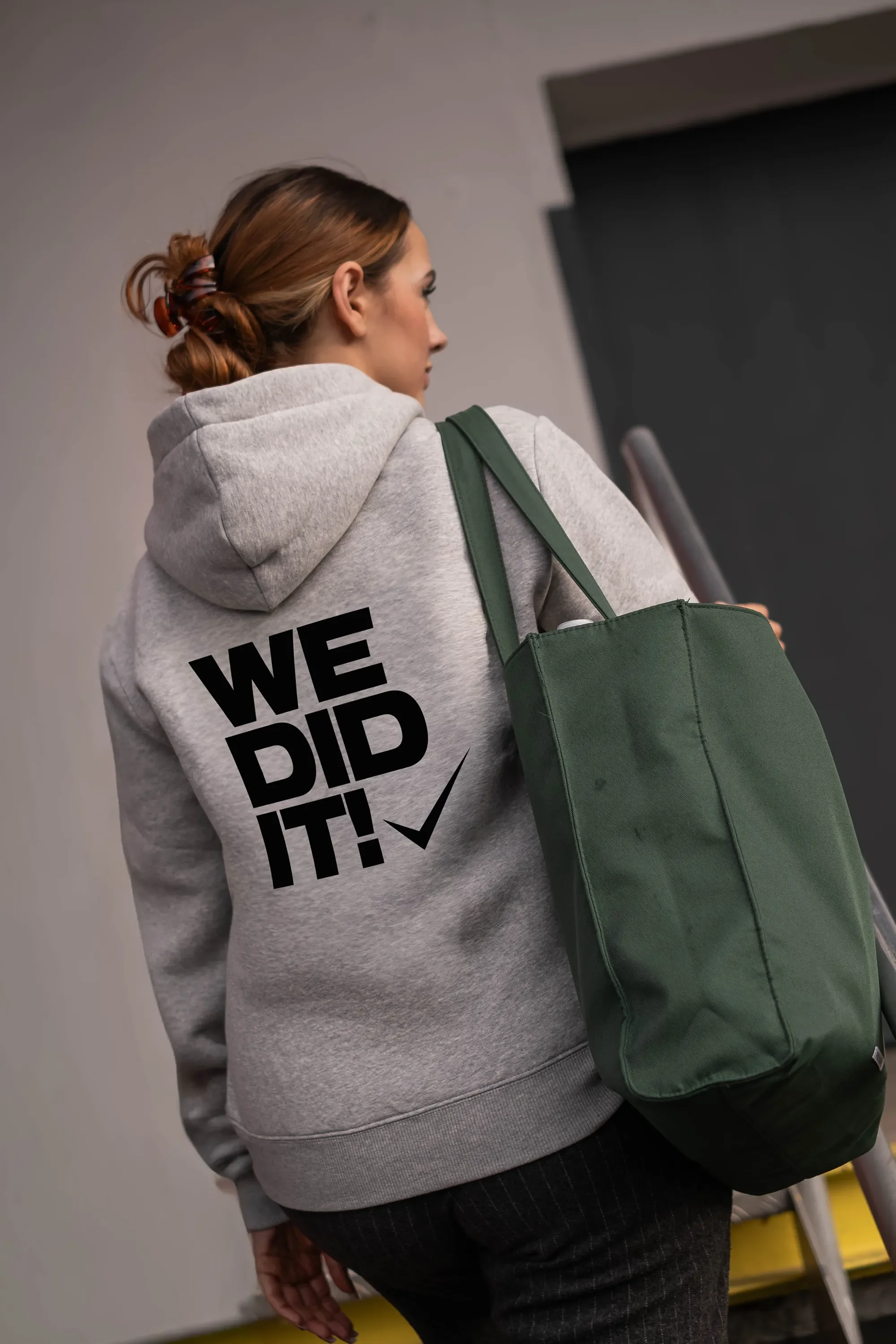 Frau mit grauem Hoodie, auf dem ‚We Did It!‘ steht, trägt eine große grüne Tasche auf der Schulter.