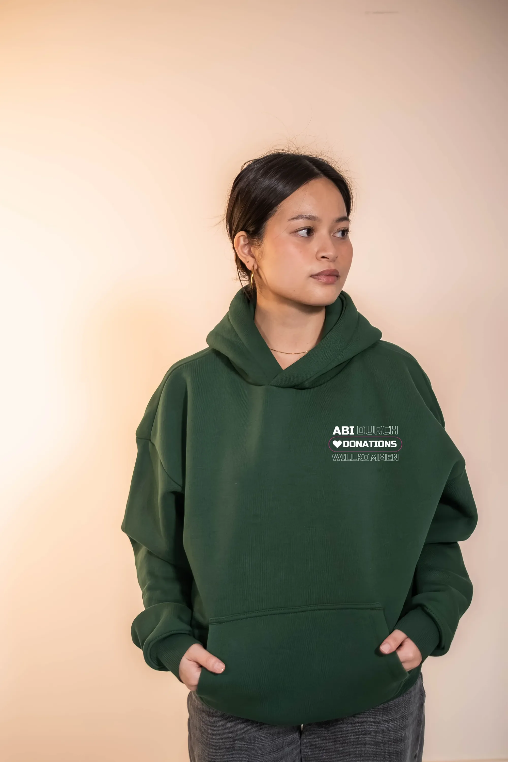 Junge Frau trägt grüne Hoodie mit text und schaut zur Seite, vor hellem Hintergrund.