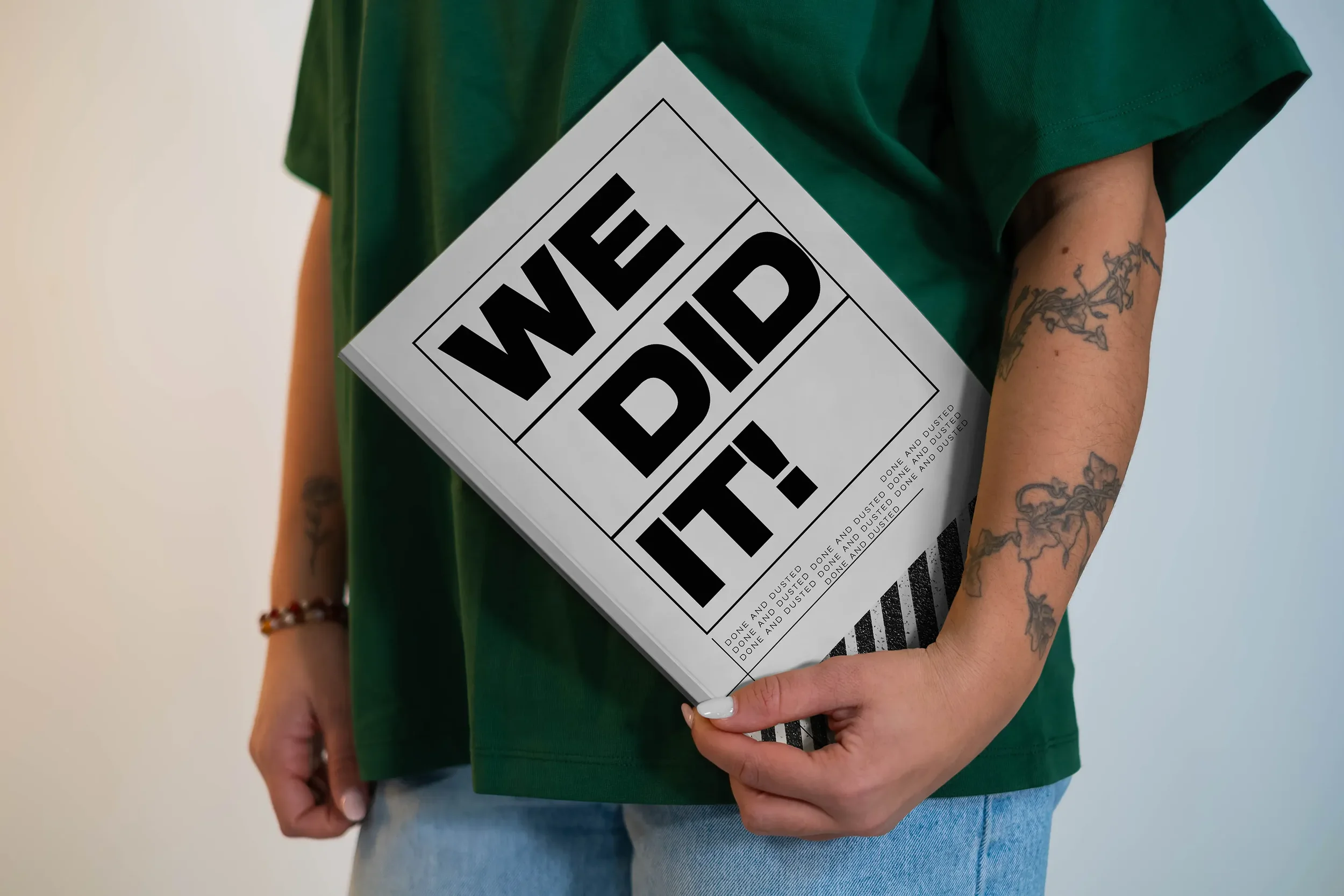 Person hält ein Buch mit dem Titel 'WEDIDIT!' in schwarz auf weißem Hintergrund. Person trägt eine grüne Kleidung, hat Tattoos am Arm, und trägt eine Armband.