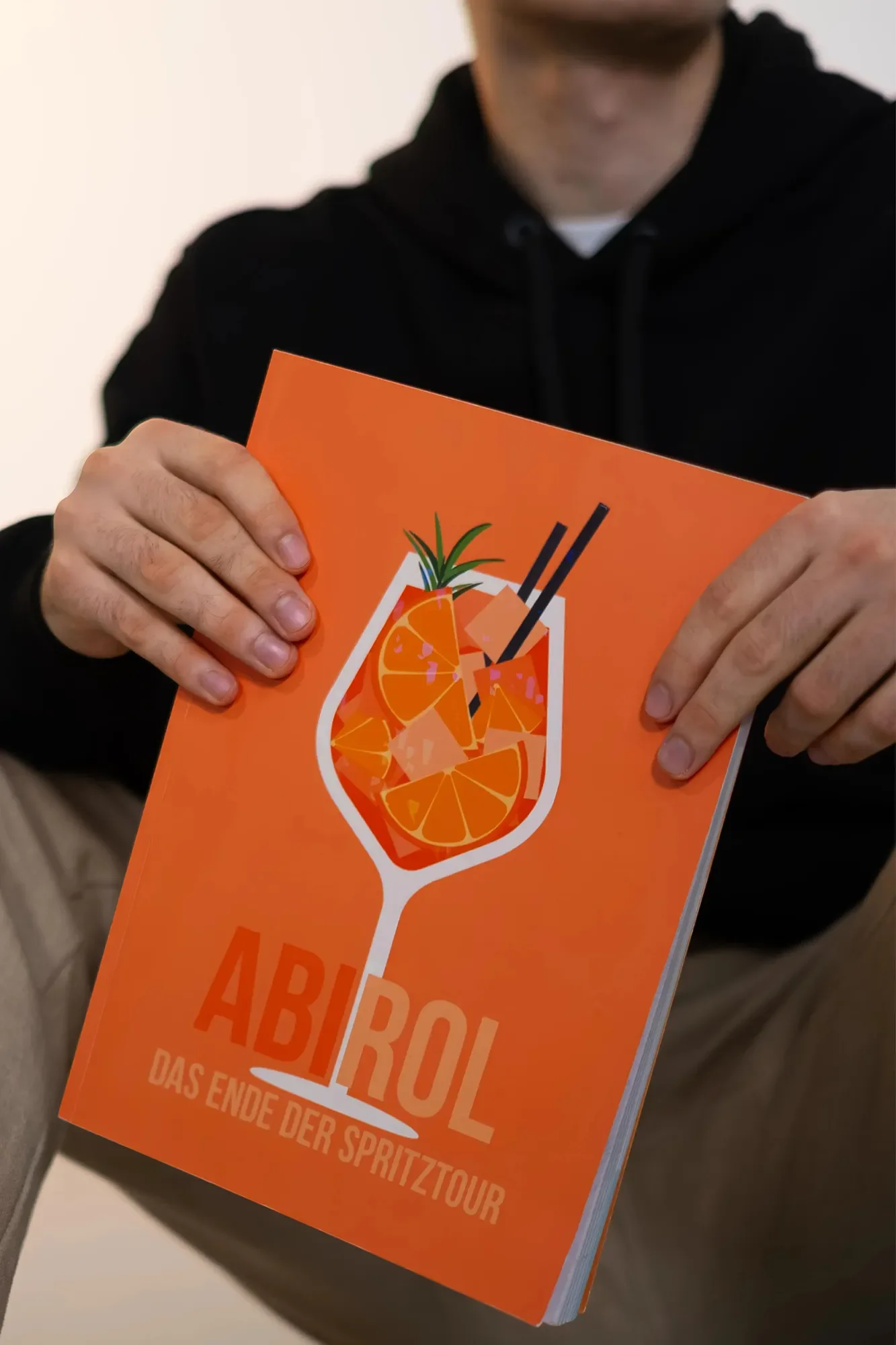 Person hält ein orangefarbenes Programm mit einem Cocktail-Design auf dem Cover, auf dem Orangen und eine Ananas zu sehen sind. Der Text lautet 'AB ROL' und 'Das Ende der Spritztour'.