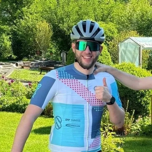 Ein lachender Mann trägt einen Fahrradhelm, Sonnenbrille und ein Fahrradtrikot, steht im Garten mit grünen Bäumen, plants und einem Gewächshaus im Hintergrund.