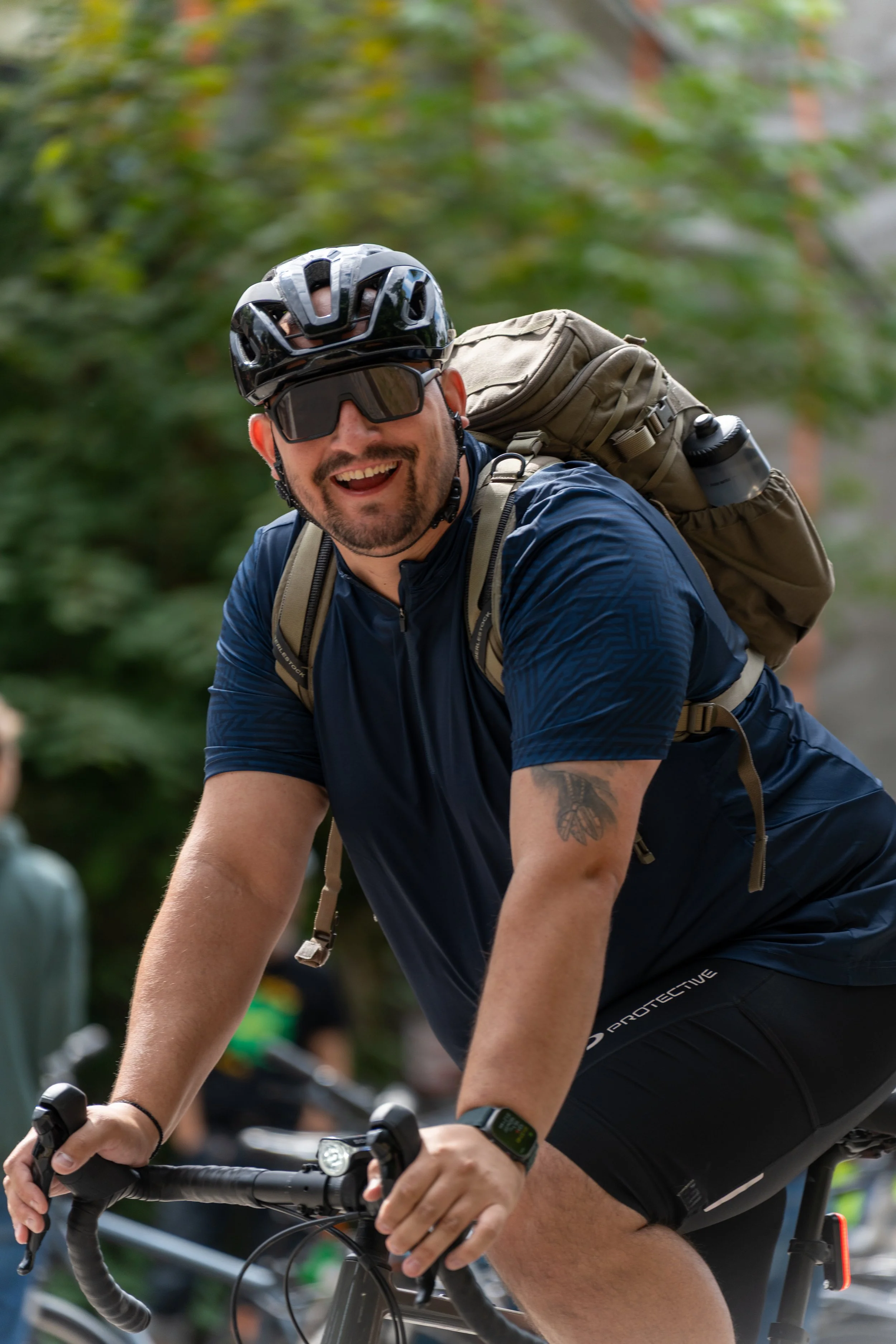 Ein lachender Mann fährt Fahrrad, trägt einen Helm und Sonnenbrille, hat einen Rucksack auf dem Rücken und ist in einer grünen, bewaldeten Umgebung unterwegs.