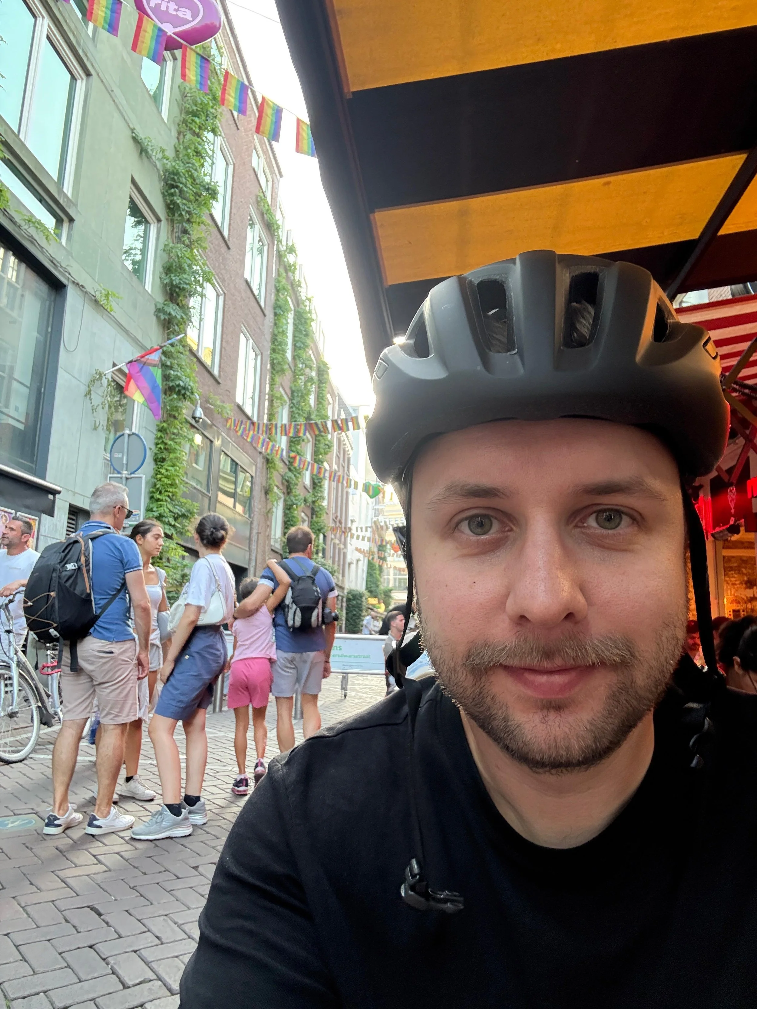 Nahaufnahme eines Mannes mit Fahrradhelm, im Hintergrund eine Menschenmenge auf einer Stadtstraße, bunt geschmückte Flaggen und grüne Pflanzen an den Gebäuden.