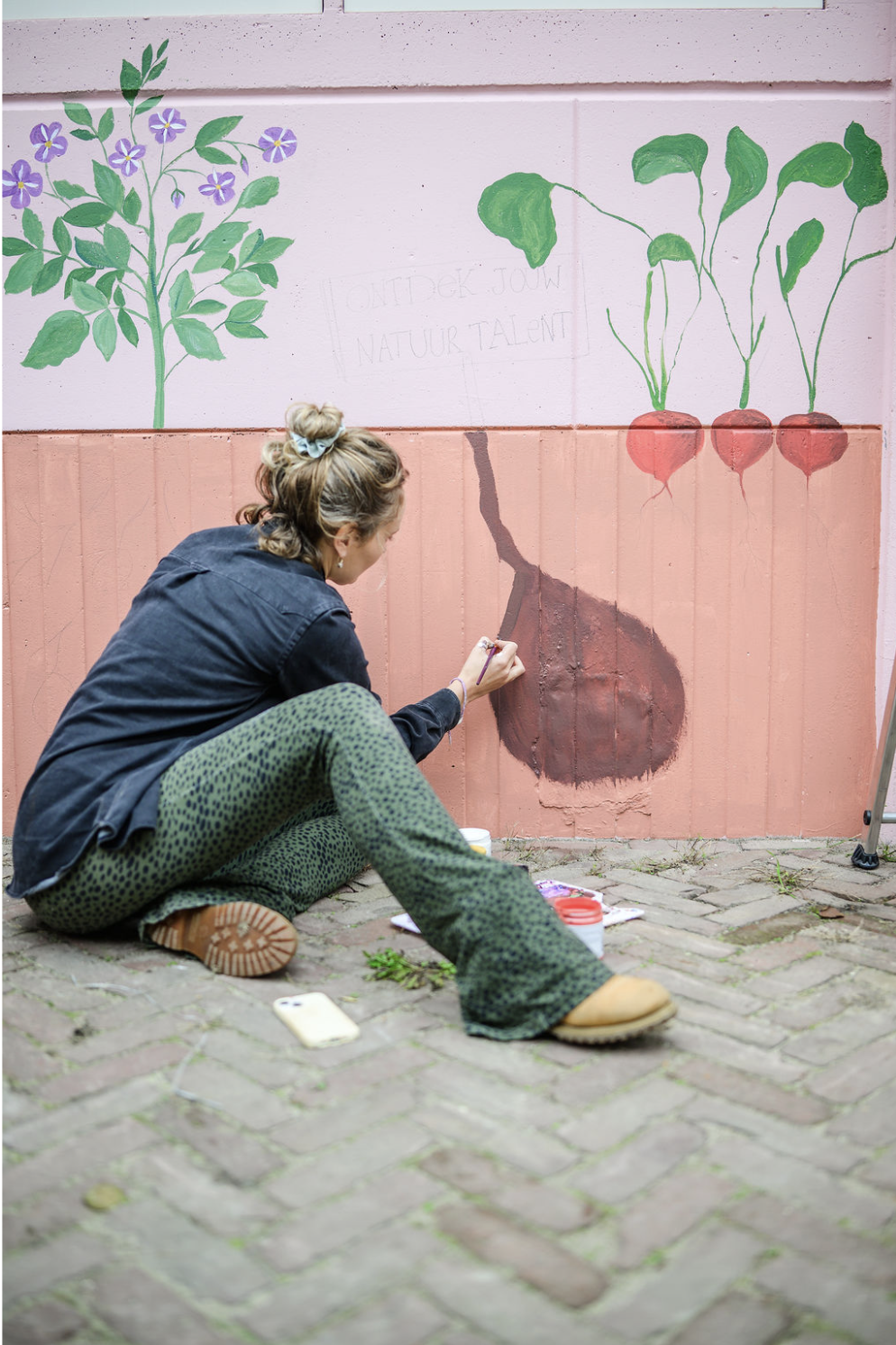 Woman sitting on brick sidewalk painting a mural of a sweet potato with green leaves and another plant with radishes on a pink wall.