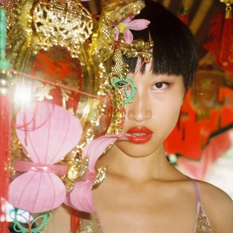 A woman with short black hair and bright red lipstick stands behind a decorative golden ornament adorned with pink paper lanterns. She gazes directly into the camera with a serious expression. The background is decorated with red elements, giving a festive or cultural atmosphere.