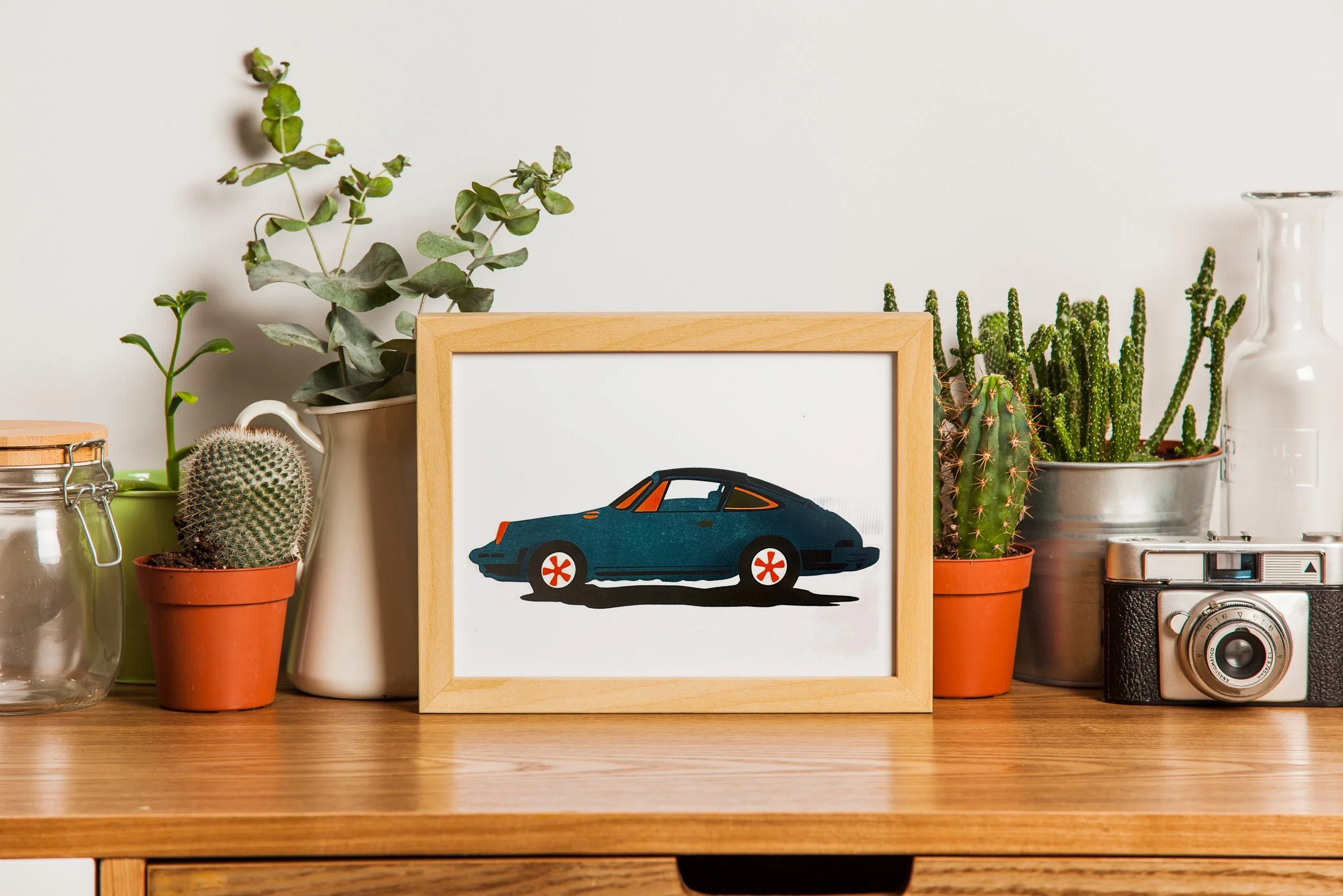 Decorative tabletop featuring framed illustration of a vintage blue car, potted succulents and cacti, glass jar, and vintage camera on wooden surface.