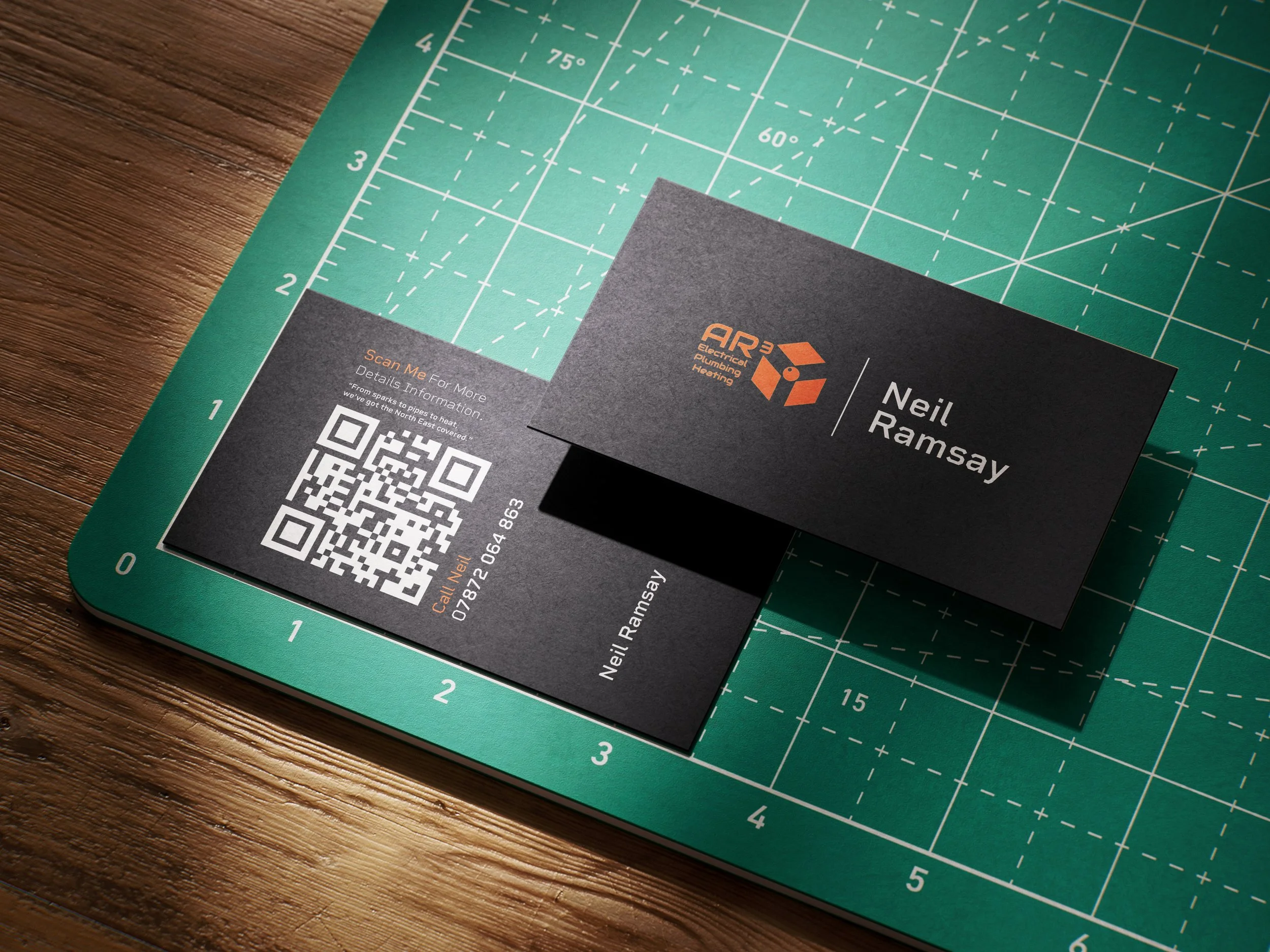 Business card for Neil Ramsay with the logo of API Electrical, Plumbing, Heating, on a business card placed on a green cutting mat with rulers, on a wooden table.