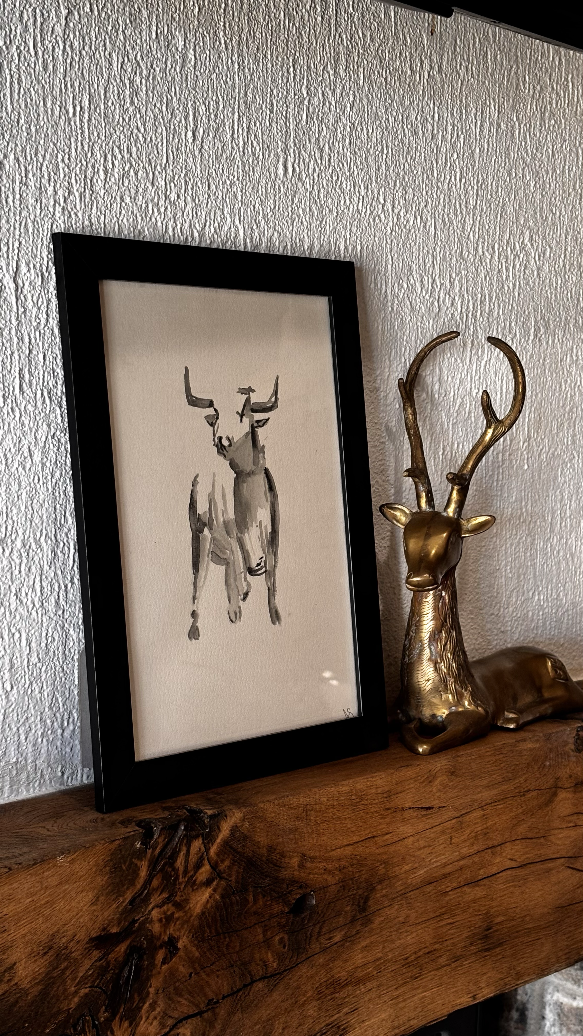 A framed watercolor painting of a deer with antlers on a textured white wall, positioned next to a gold-colored decorative deer statue on a wooden surface.