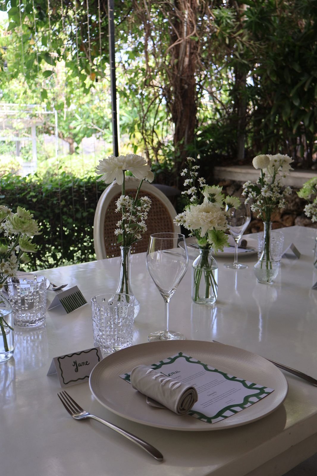 A table set for a formal event outdoors with floral centerpieces, wine glasses, water glasses, and plates, surrounded by greenery and trees. Hand-painted stationery menu and placement cards.