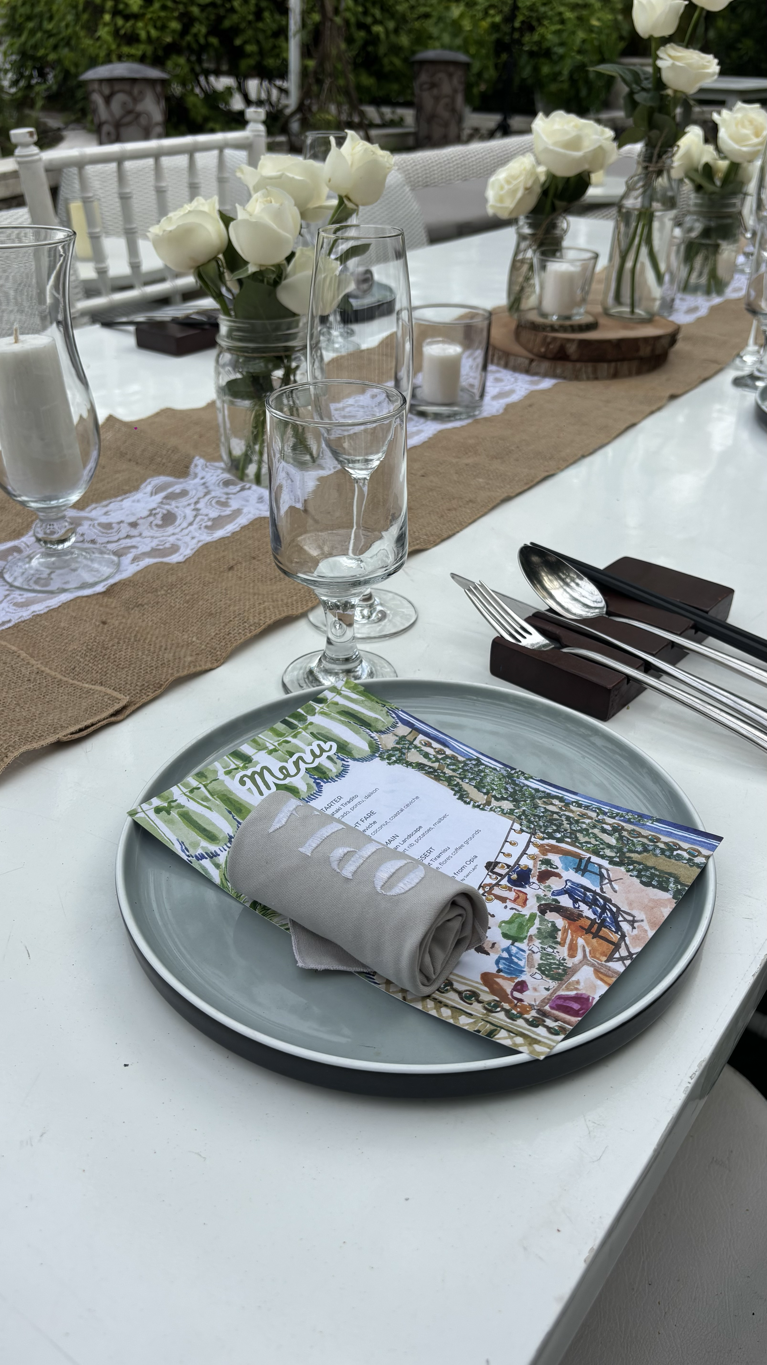 Elegant outdoor dining table setup with white roses in glass vases, candles, menu, napkin, and silverware, on a white table with a burlap table runner and hand-painted stationery.