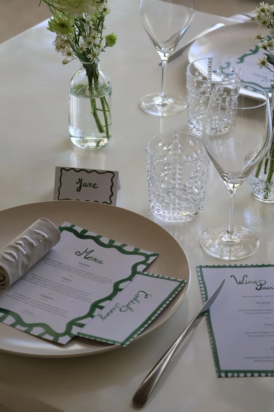 A formal dining table setting with a white tablecloth, floral centerpiece in a glass jar, wine glasses, crystal water glasses, and green hand-painted watercolor stationery collection.